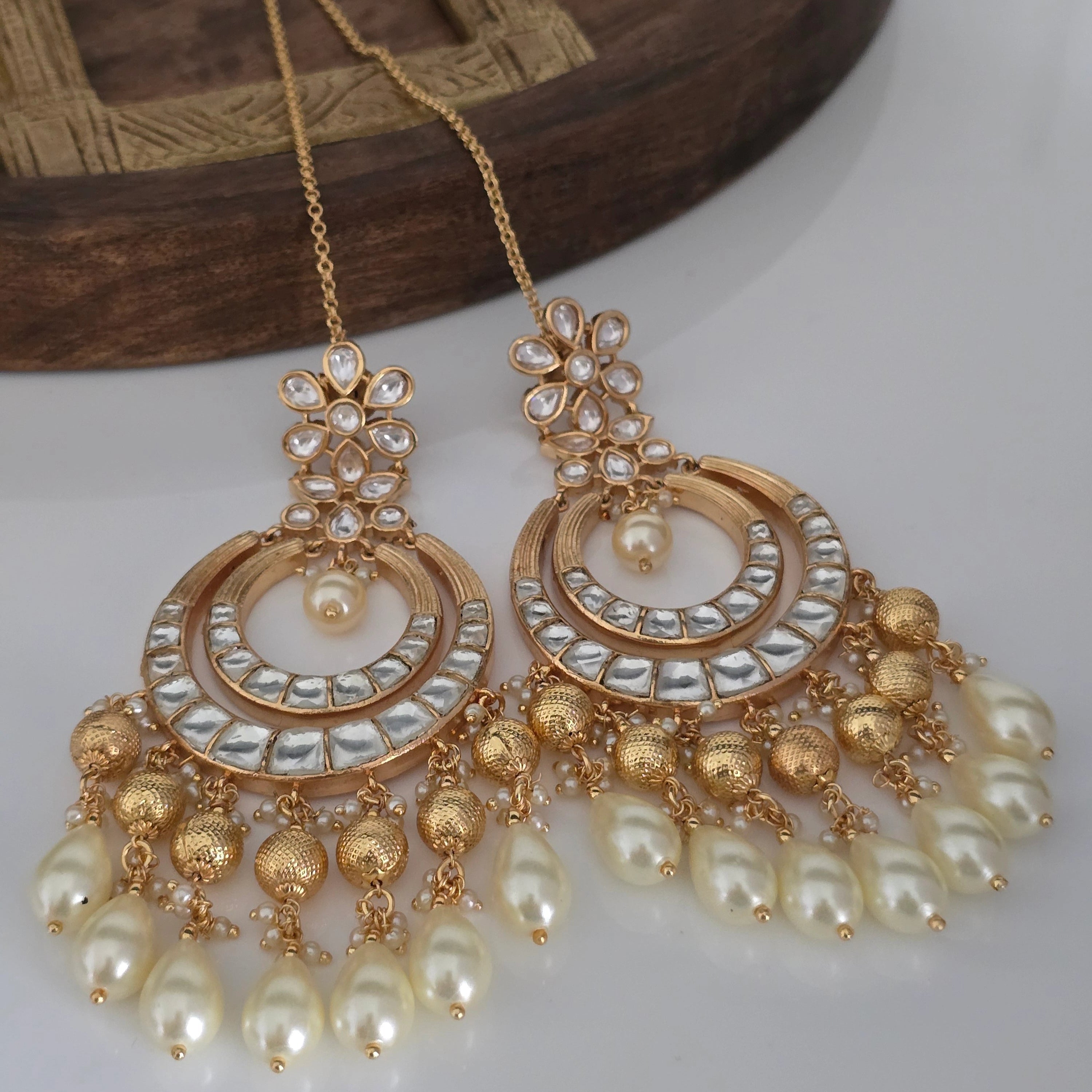 Two gold-tone Chandbali earrings featuring Kundan stones, textured gold beads, and white pearl drops, each with a delicate ear chain, presented on a light surface.