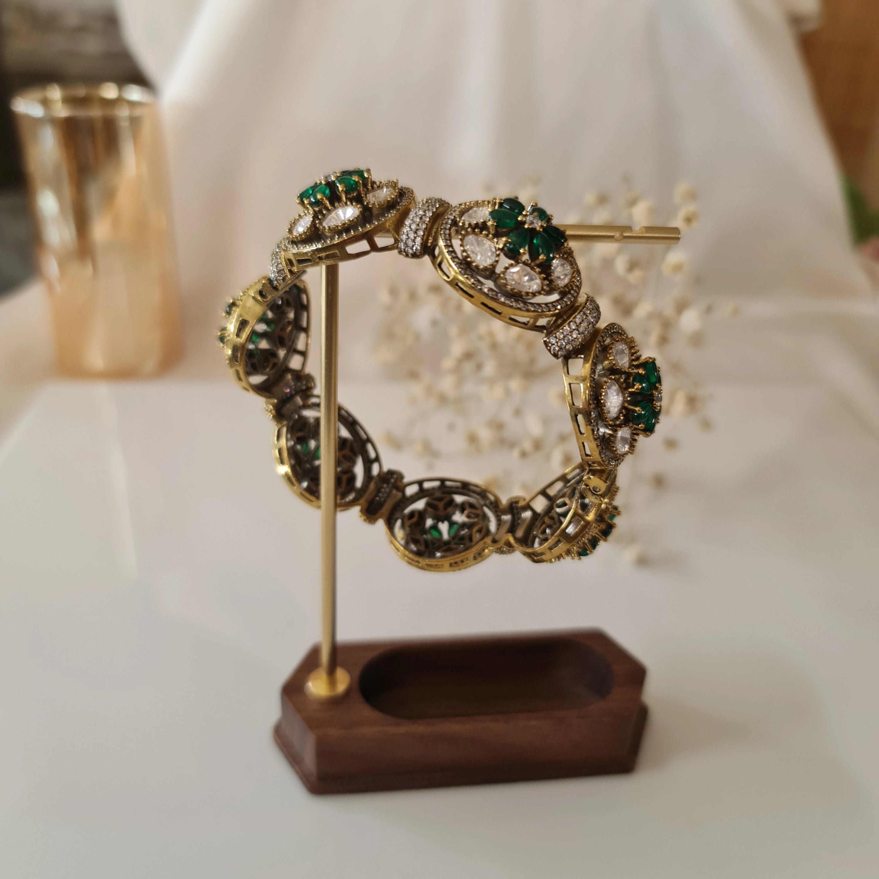 Gold-plated kada bracelet with green emerald and American diamond motifs displayed on a wooden jewelry stand another view.