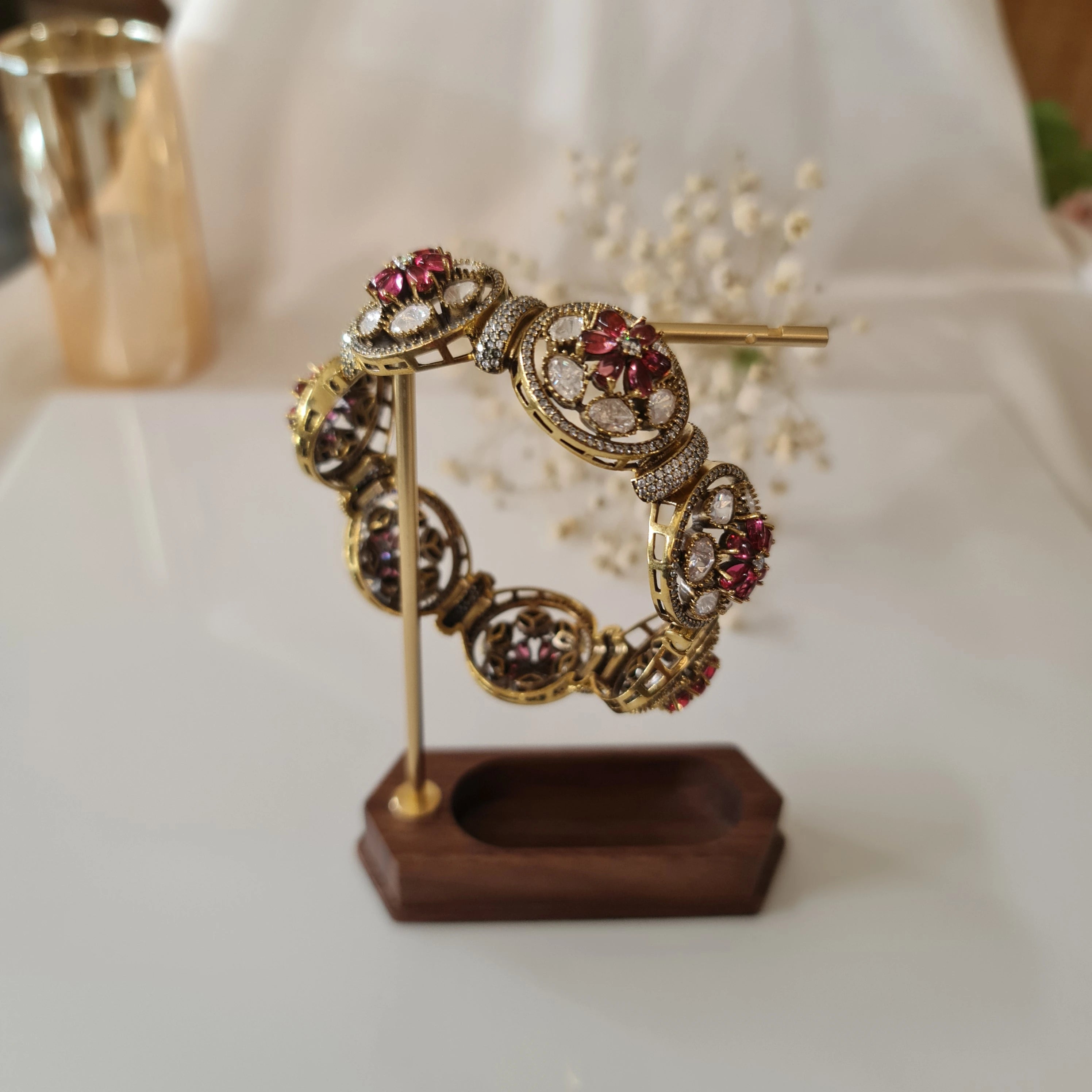 Gold-plated kada bracelet with ruby red and CZ motifs displayed on a wooden jewelry stand.