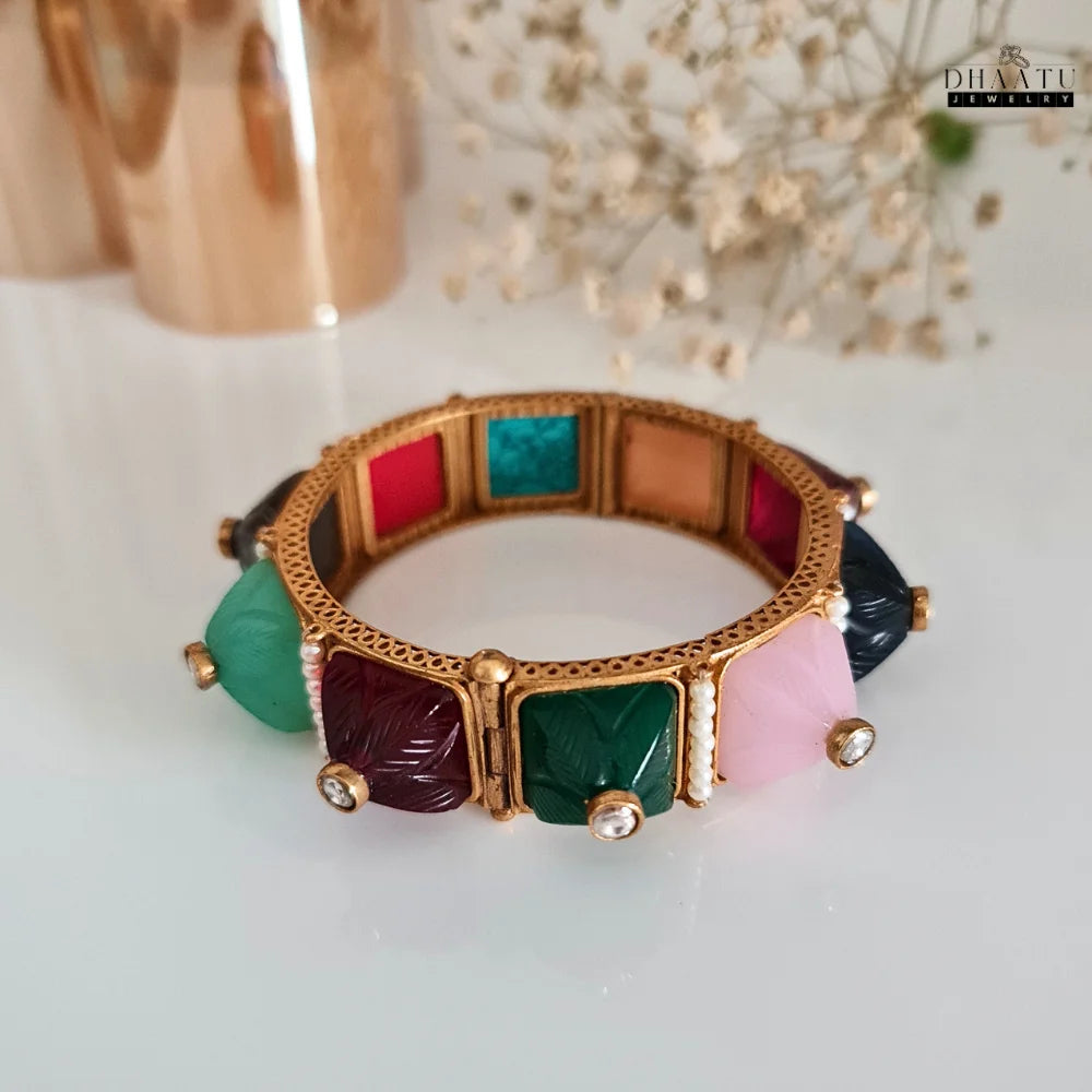 Close-up shot of a multicolor carved stone and pearl kada bracelet on a golden display stand, showing orange, maroon, black, and turquoise stones.