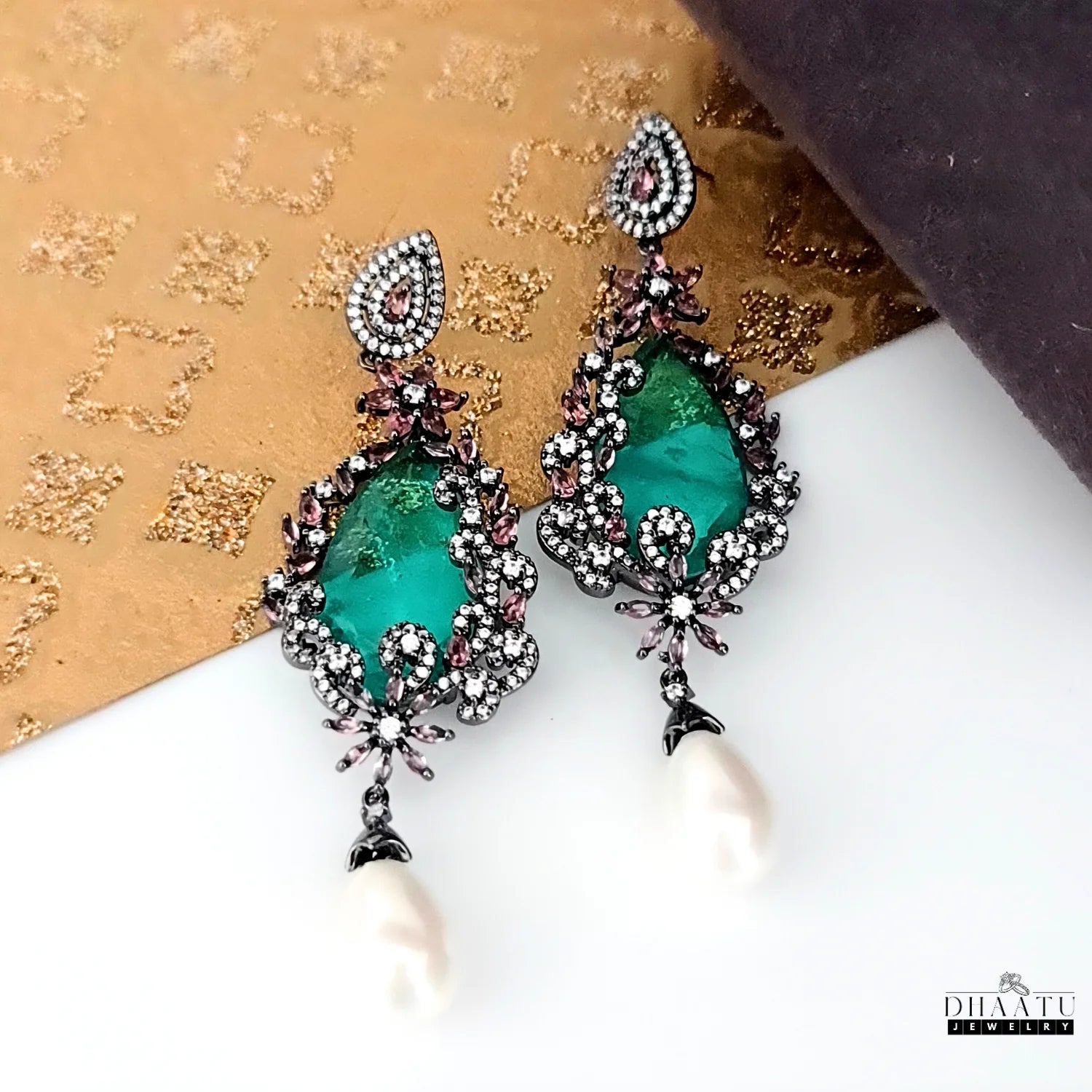 Angled view of DHAATU JEWELRY green doublet stone and pearl drop earrings with a patterned gold background.