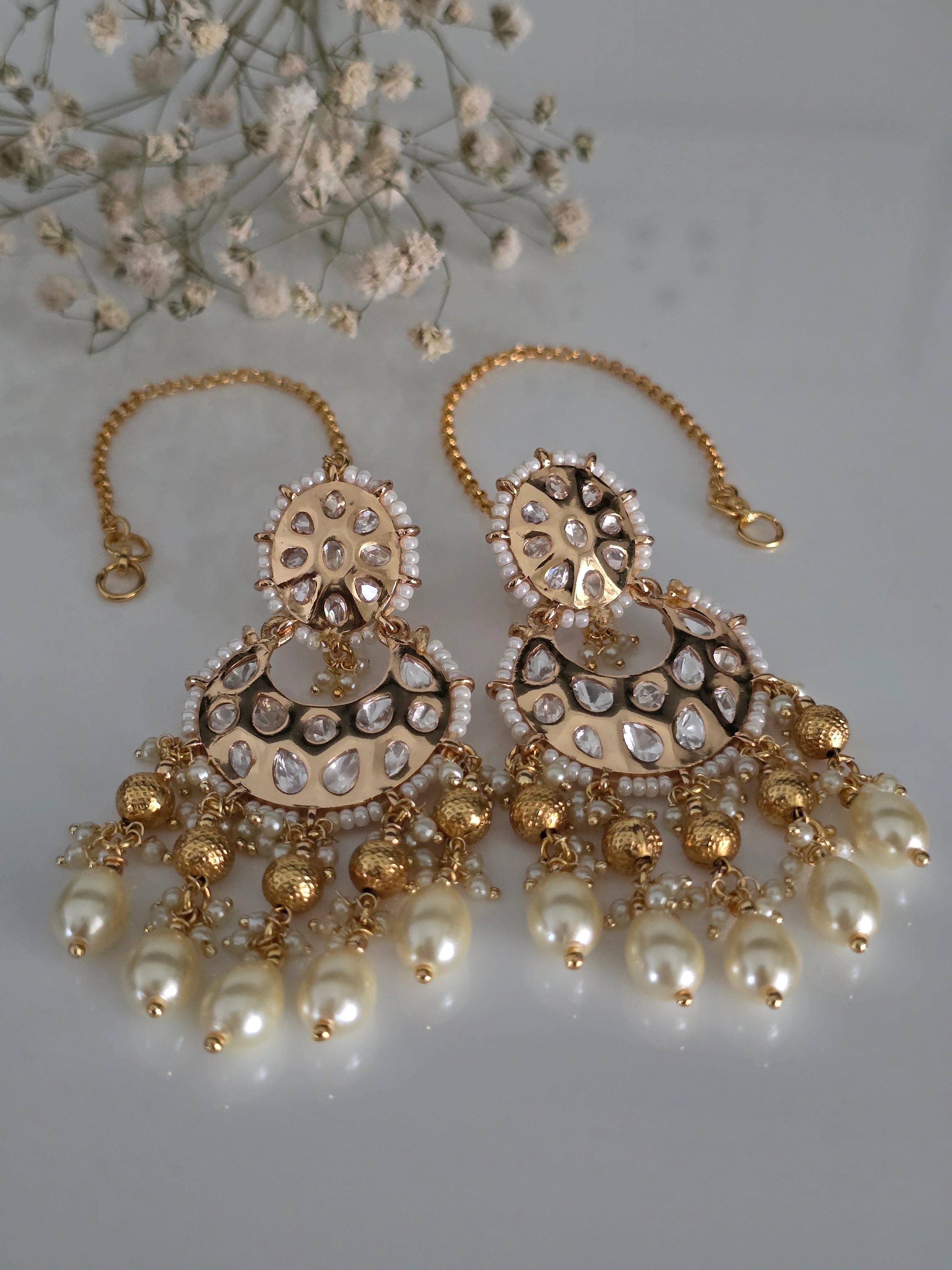 Close-up of ornate Indian bridal earrings with crescent shape, uncut look stones, small white pearls, and dark purple beads, with ear chain.