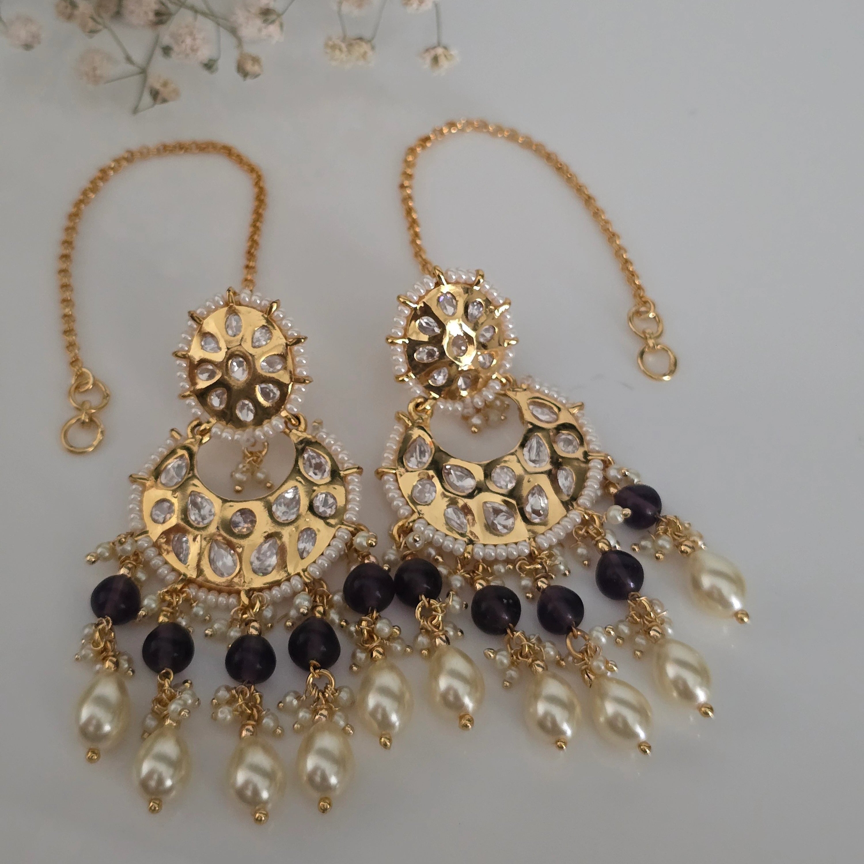 Pair of gold-tone traditional Indian Chandbali dangler earrings featuring Polki-style American Diamonds, pearls, and purple accent beads, with an attached Sahara chain.