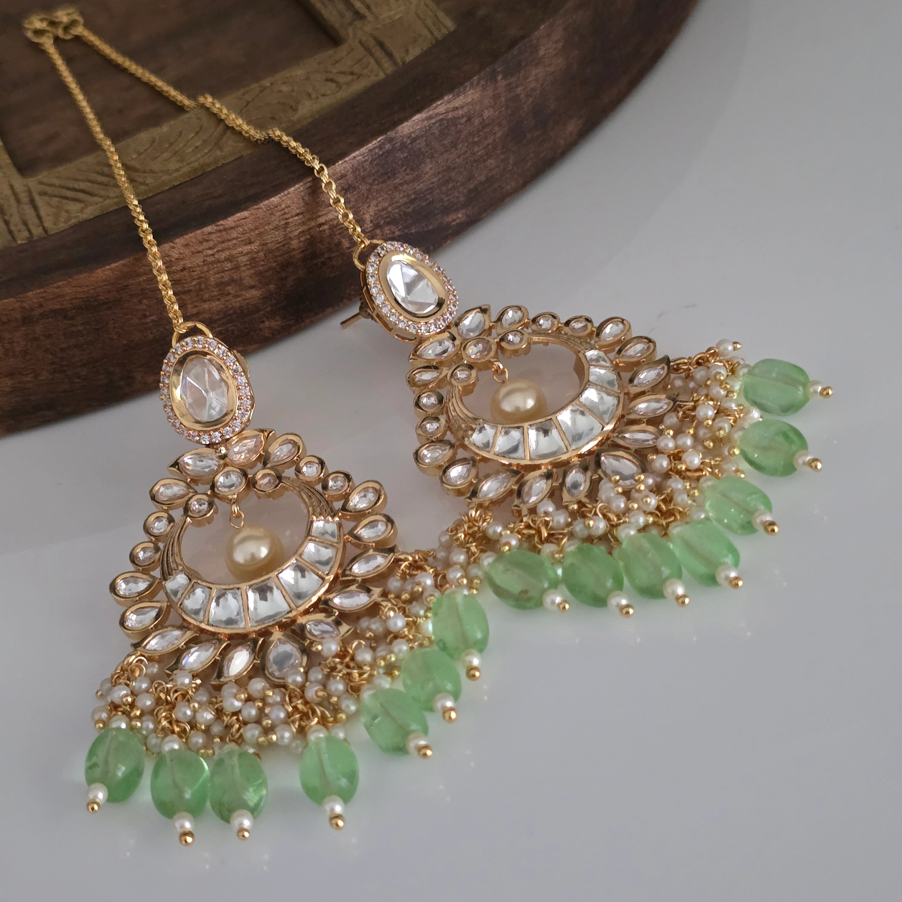 Pair of gold-tone traditional Indian Chandbali dangler earrings with Kundan, Cubic Zirconia, pearls, and light green teardrop beads, with an attached Sahara chain on a wooden and white background.