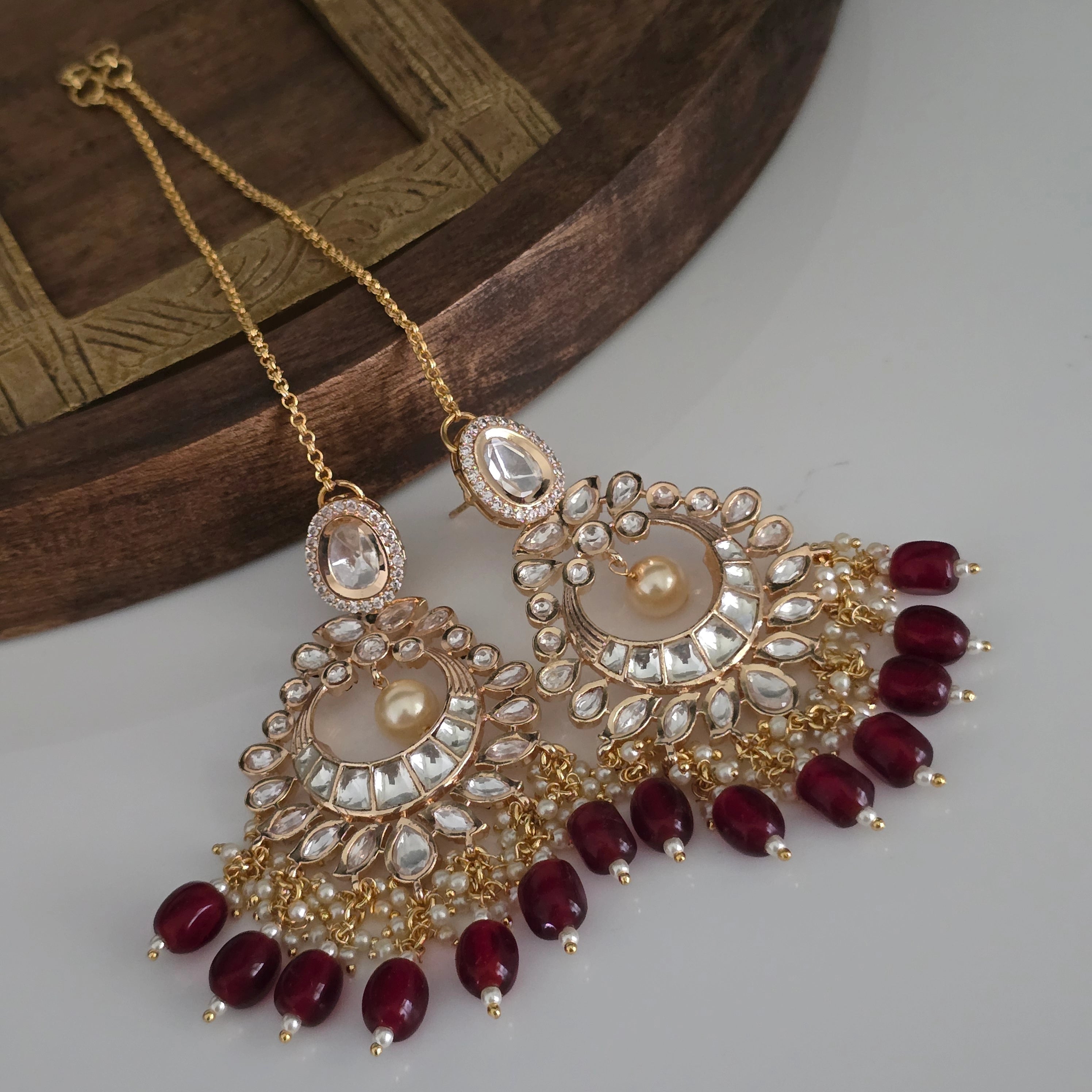 Close-up of ornate Indian bridal earrings with crescent shape, uncut look clear stones, small white pearls, and dark red beads, with ear chain on white and wood background.