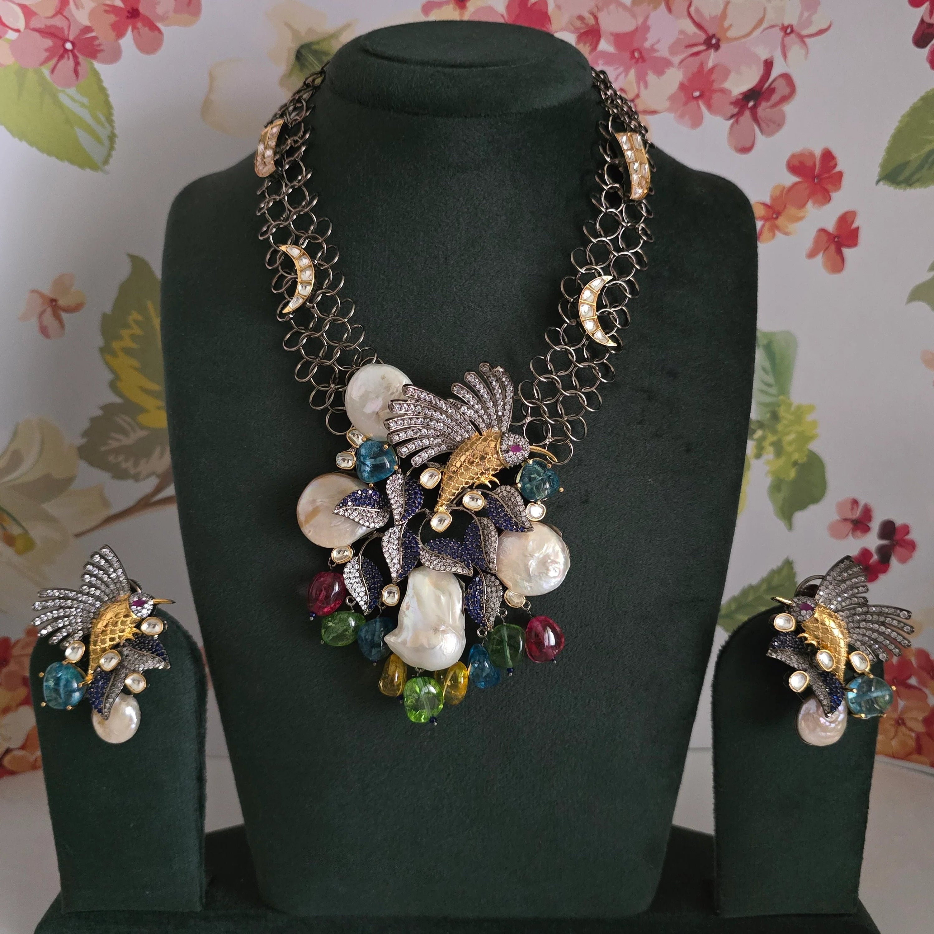 Complete artificial jewelry set, including the hummingbird statement necklace and matching earrings, displayed on a green jewelry bust against a floral backdrop.