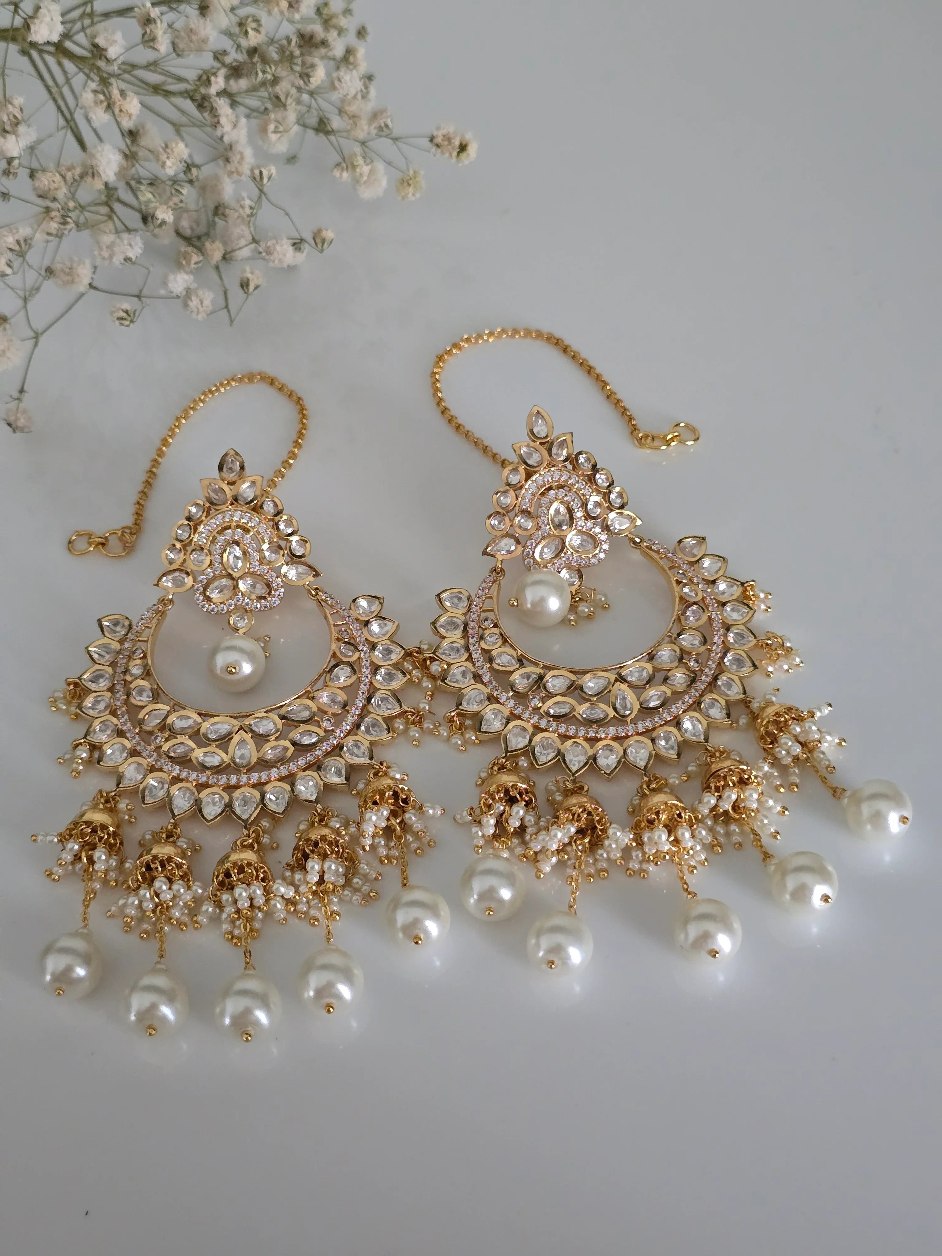 Gold Plated Kundan and Pearl Chandbali Earrings with delicate jhumka drops and ear chains, artfully arranged with white gypsophila flowers, highlighting their elegant and festive appeal.