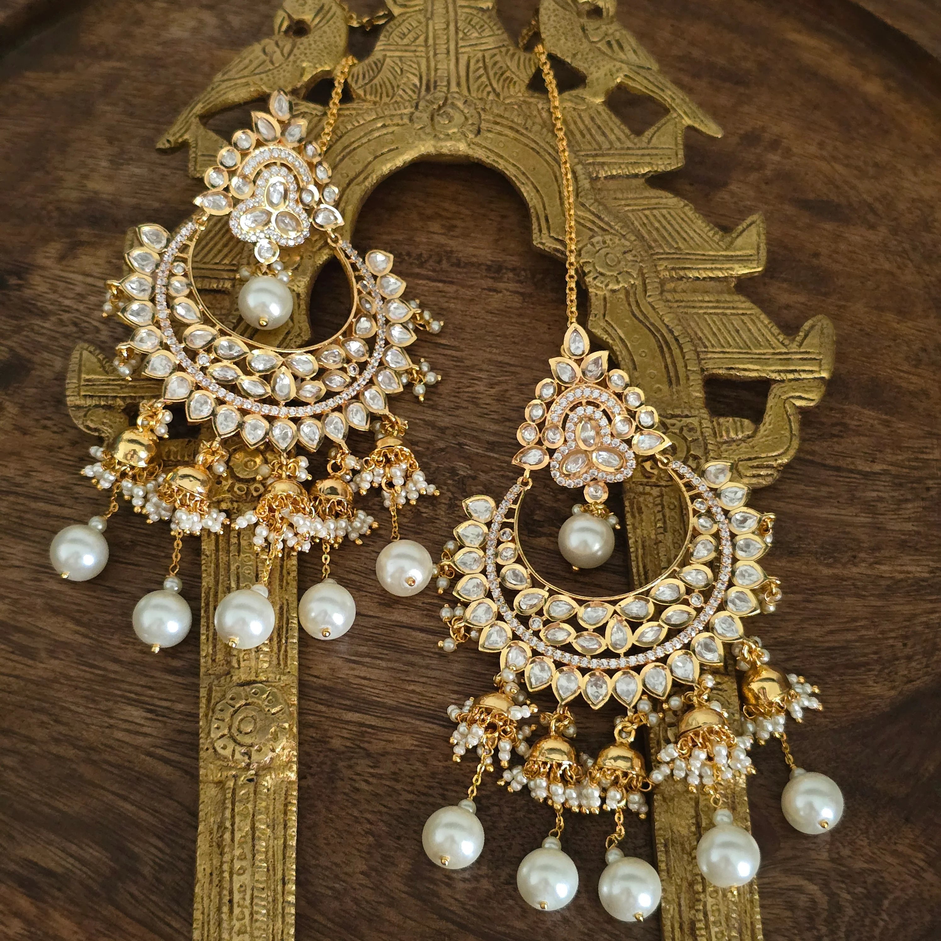 Stunning Gold Plated Kundan and Pearl Chandbali Earrings with attached ear chains displayed on a traditional wooden background, showcasing intricate details and jhumka drops.