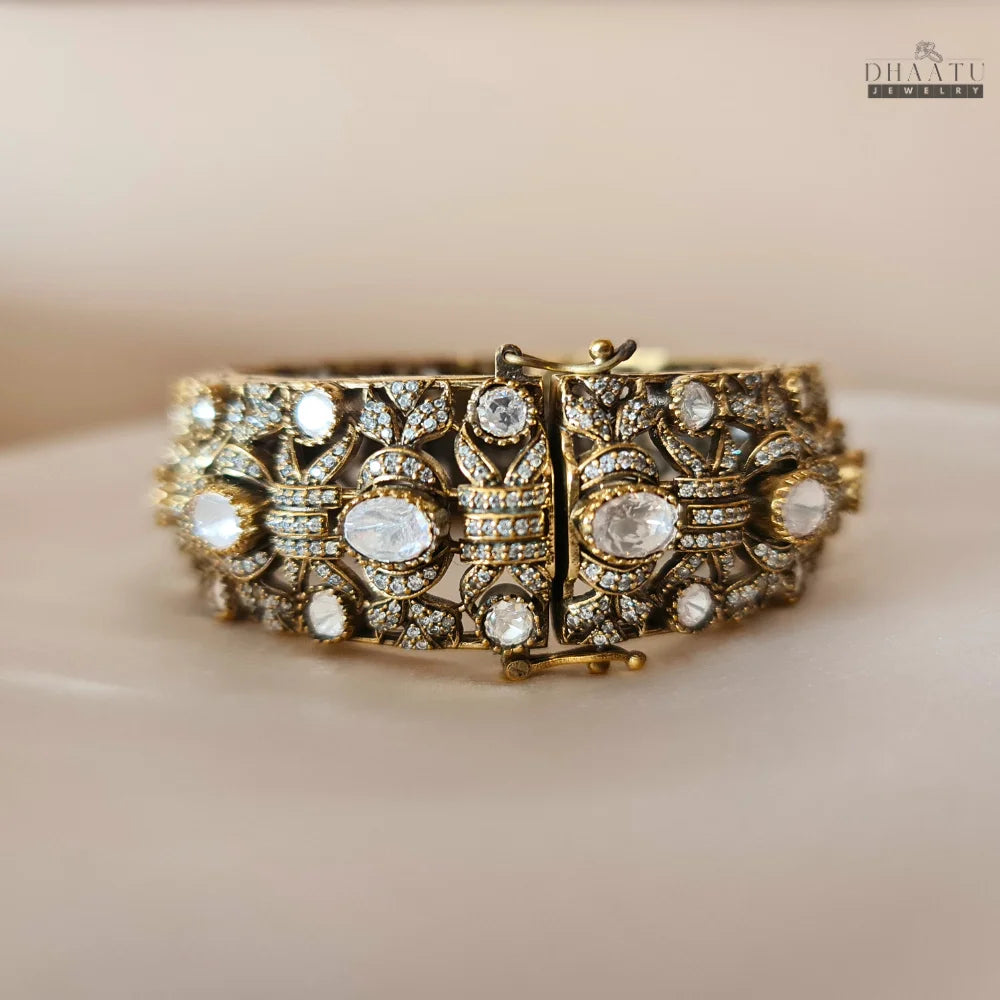 Front view of a gold-plated kada bracelet with polki diamond style stones and intricate detailing, on a white surface with pink flowers in the background.
