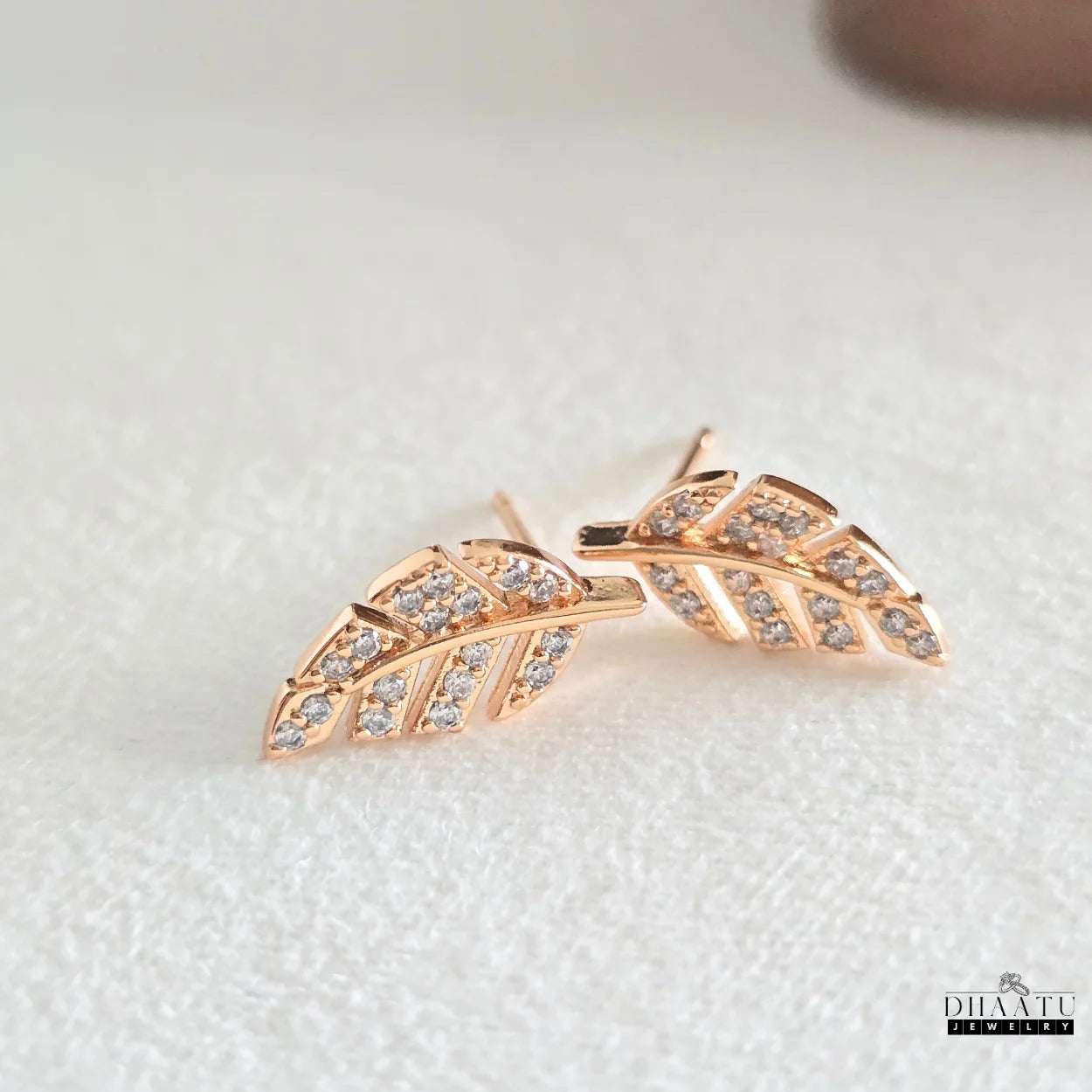 Pair of Dhaatu Jewelry rose gold-plated leaf stud earrings featuring brilliant zirconia, lying on a white textured surface.

