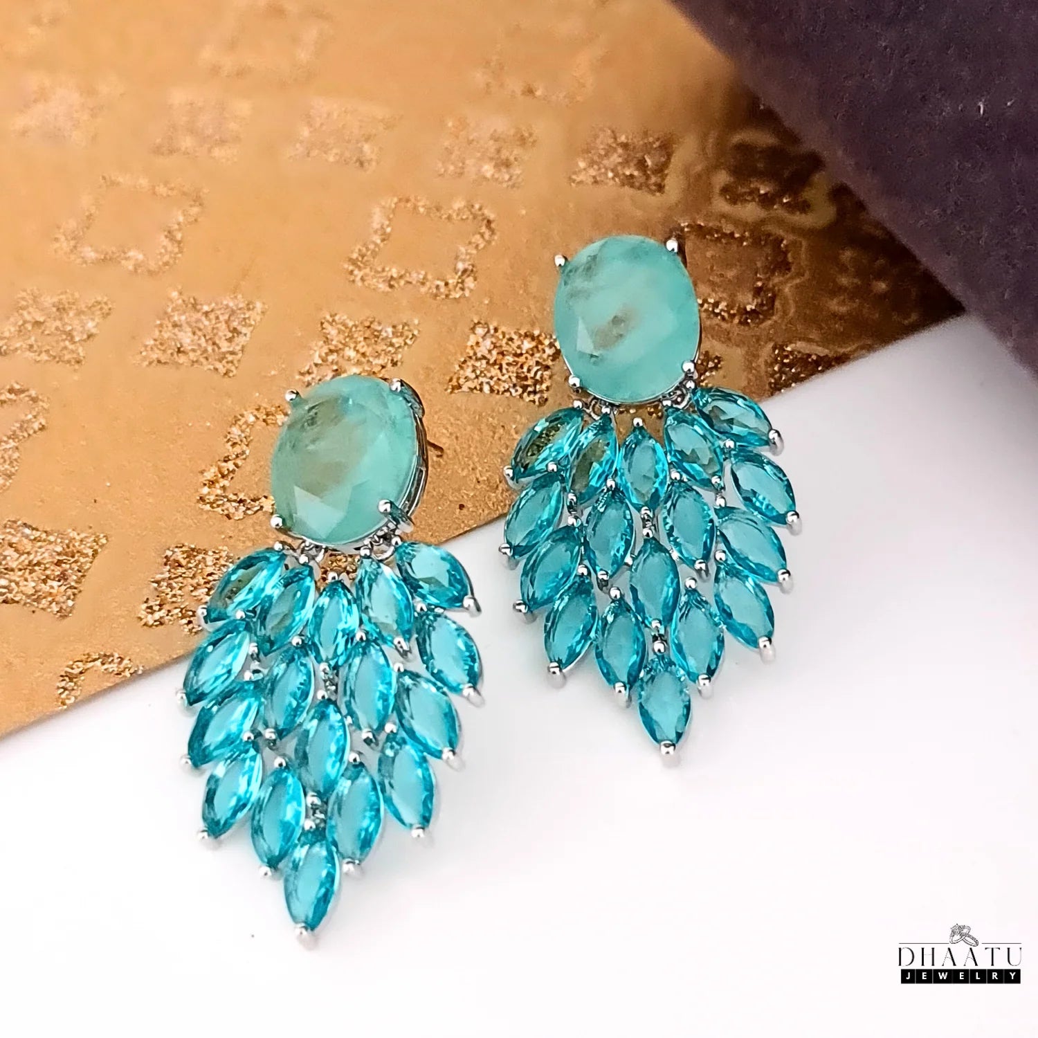 A close-up shot of the stunning Aqua Chalcedony and marquise cut blue stone dangler earrings, showcasing the intricate details and craftsmanship on a textured gold and white background.
