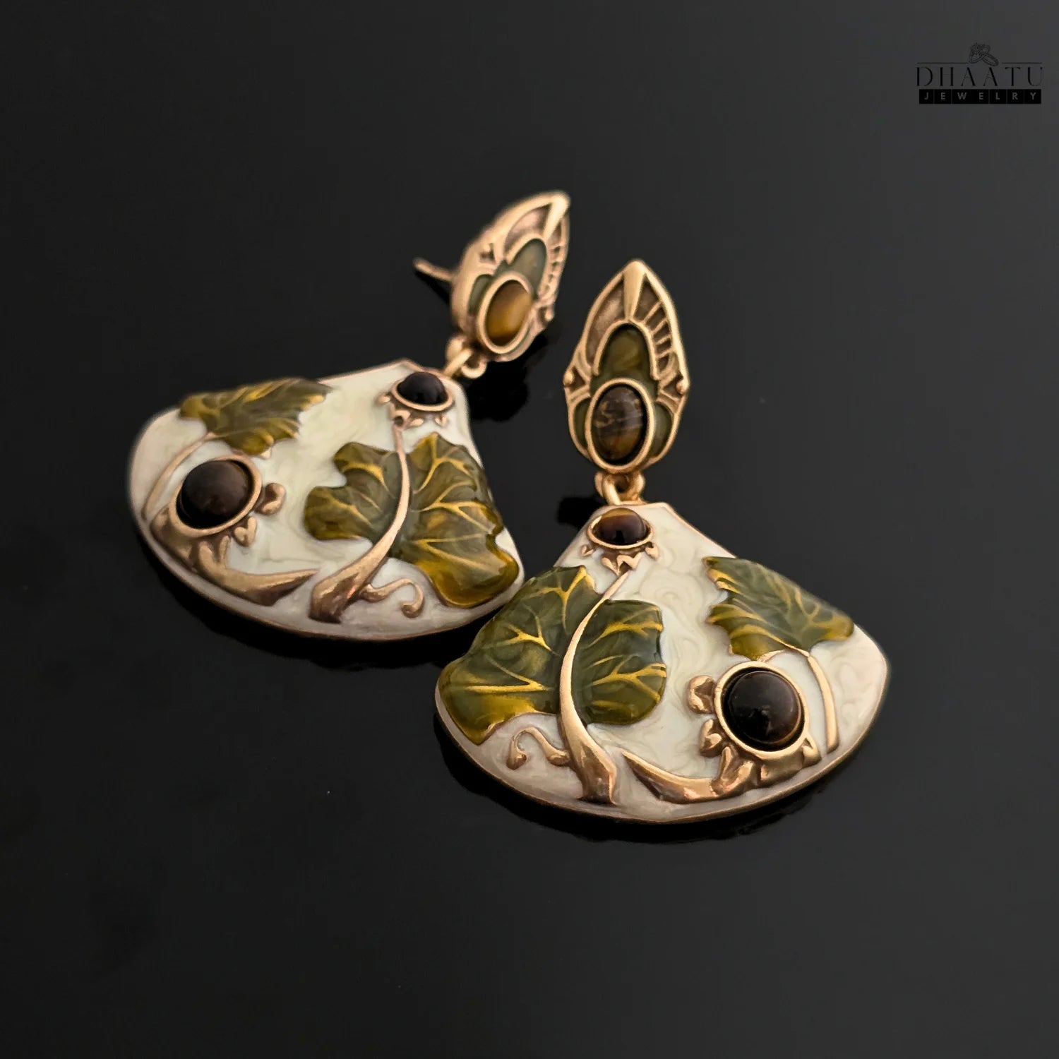 A pair of Art Nouveau lotus drop earrings with white enamel and green leaves, set against a reflective black background.