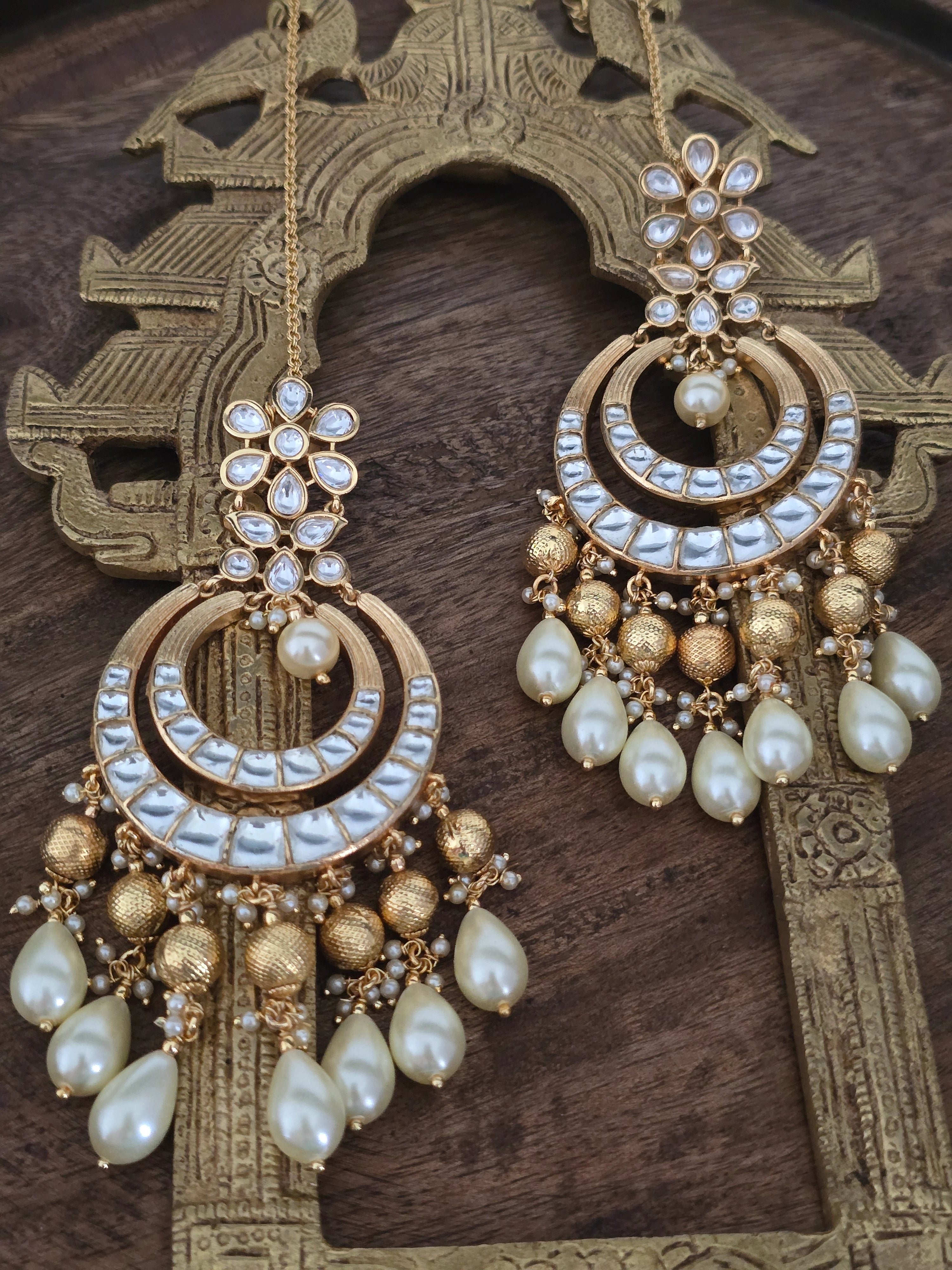 Pair of ornate gold-tone Chandbali dangler earrings with Polki-style Kundan, textured gold beads, and teardrop pearls, with an attached Sahara chain, displayed on a wooden stand.