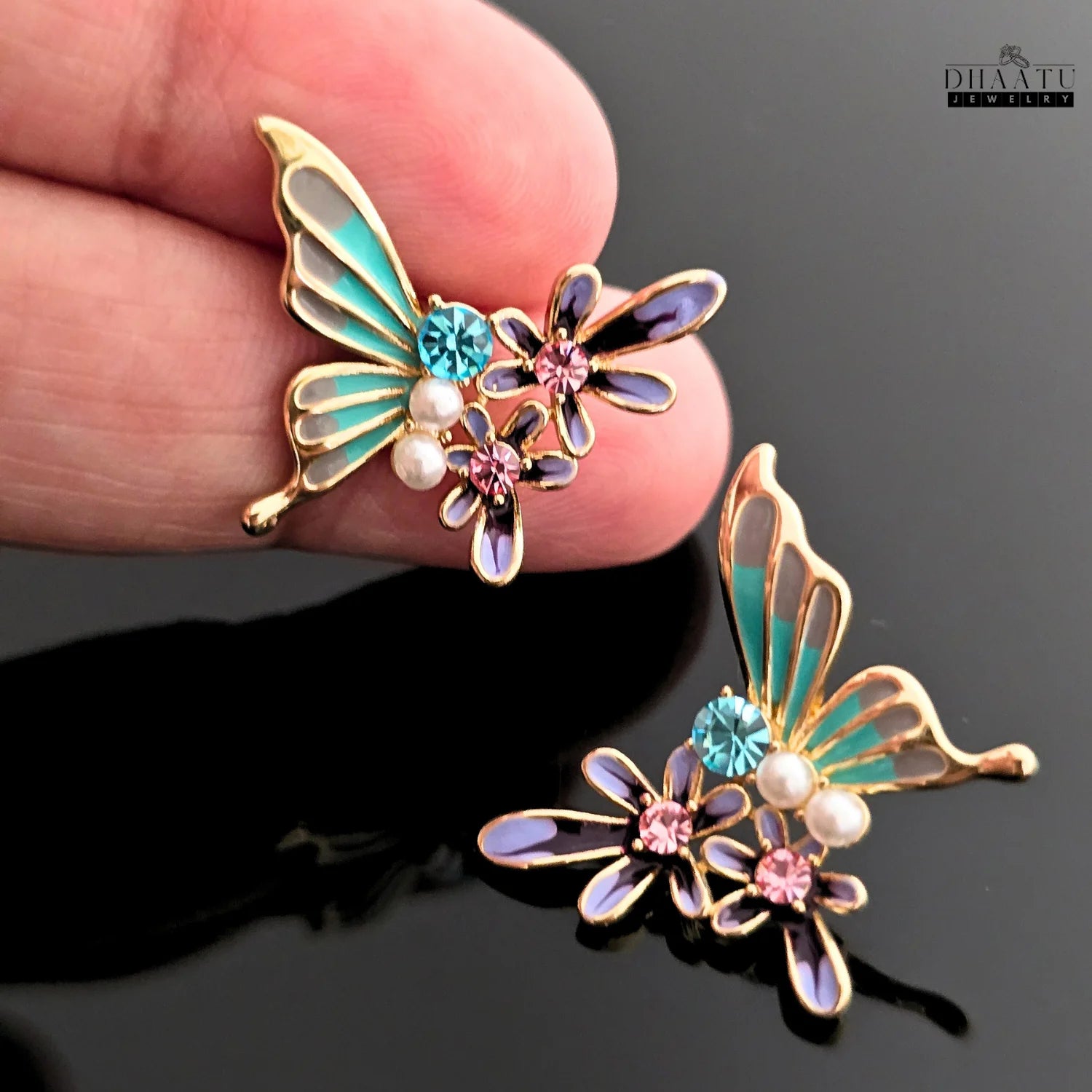 Close-up of a hand holding a turquoise and purple butterfly and flower earring to show scale.