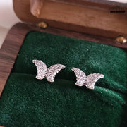 Pair of glittering butterfly stud earrings in silver tone with zircon embellishments, displayed on green velvet in a wooden jewelry box.