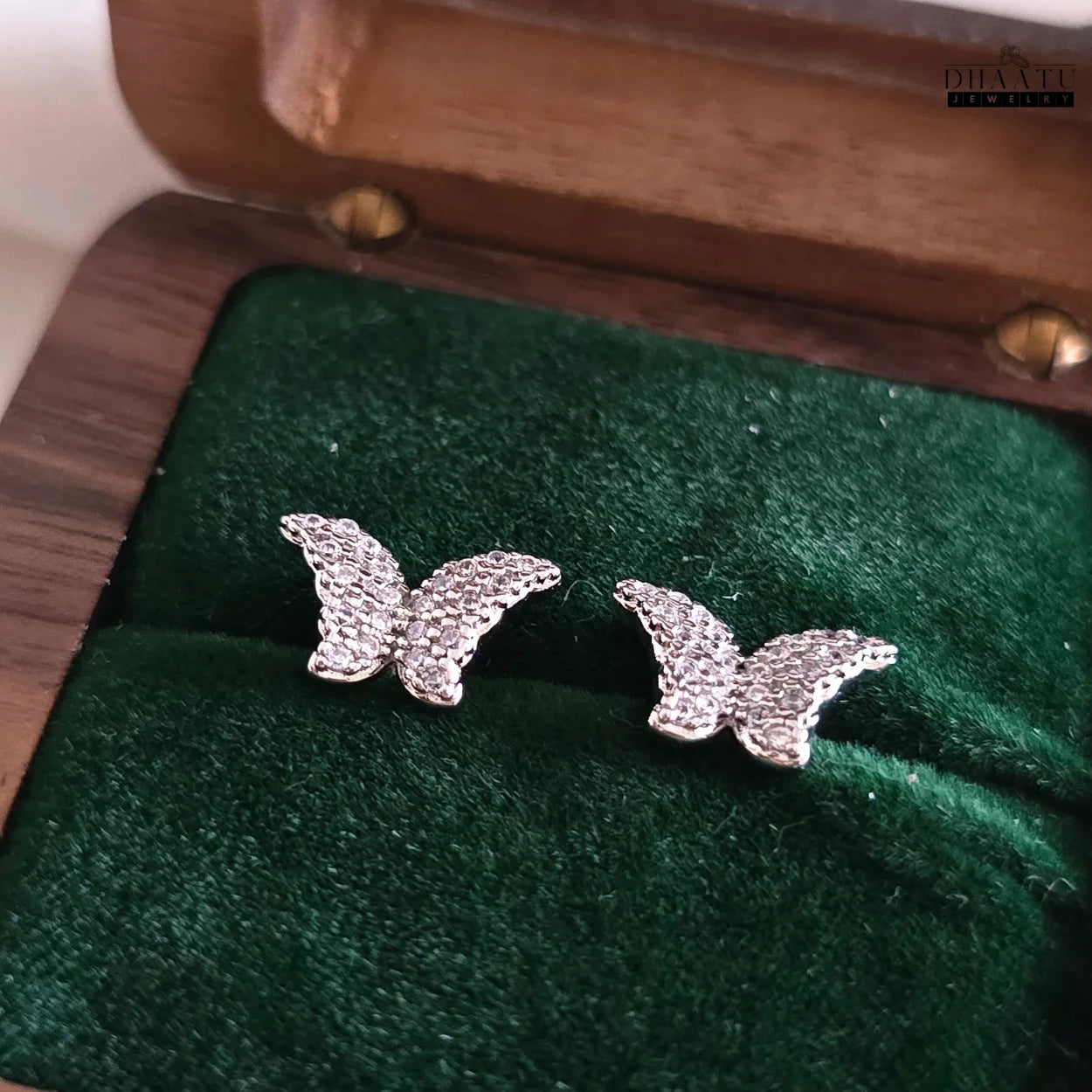 Pair of glittering butterfly stud earrings in silver tone with zircon embellishments, displayed on green velvet in a wooden jewelry box.