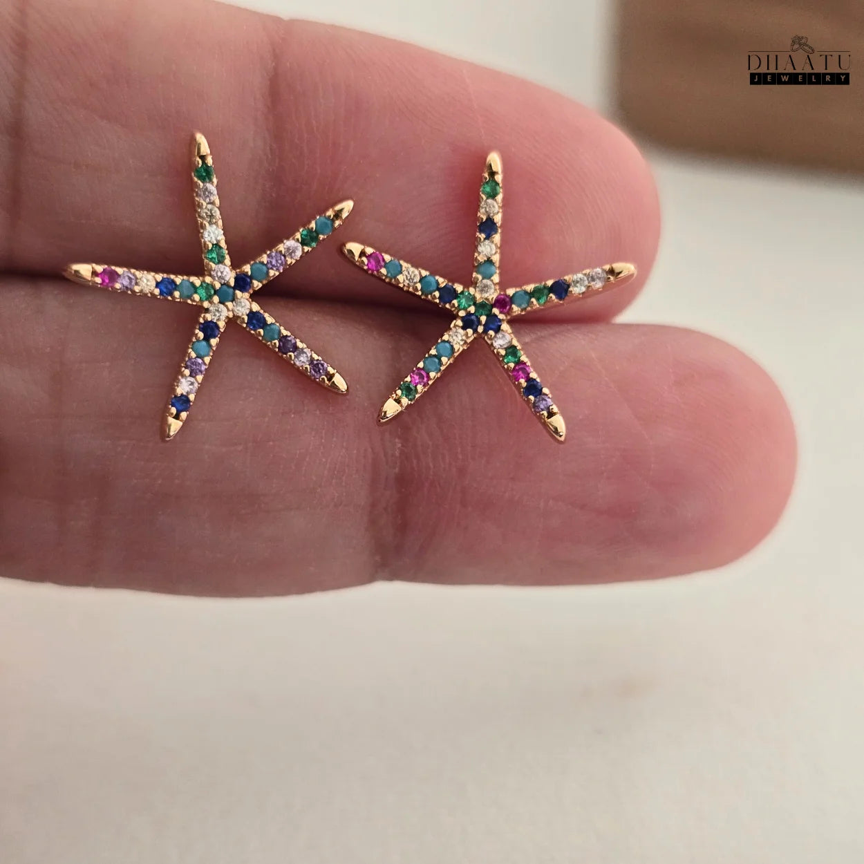 Detailed close-up of two rose gold starfish stud earrings with a medley of colorful zircon stones, held delicately on a person's fingers to show size and detail.