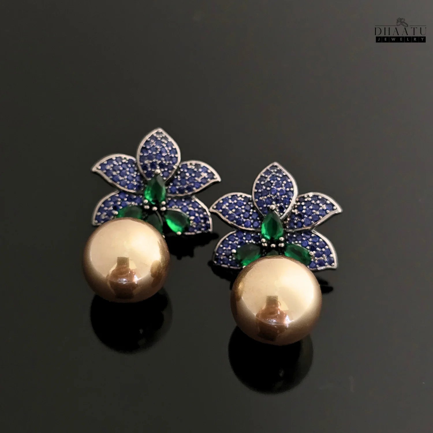 A pair of Dhaatu Jewelry blue sapphire and emerald flower earrings with pearl drops displayed on a reflective black surface.
