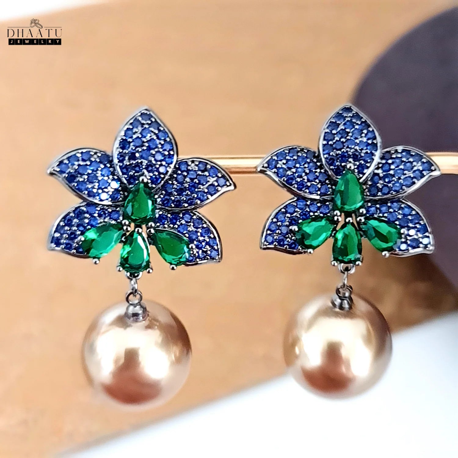 The Dhaatu Jewelry blue sapphire and emerald flower earrings with pearl drops hanging from a display bar, showcasing the length and movement of the design.