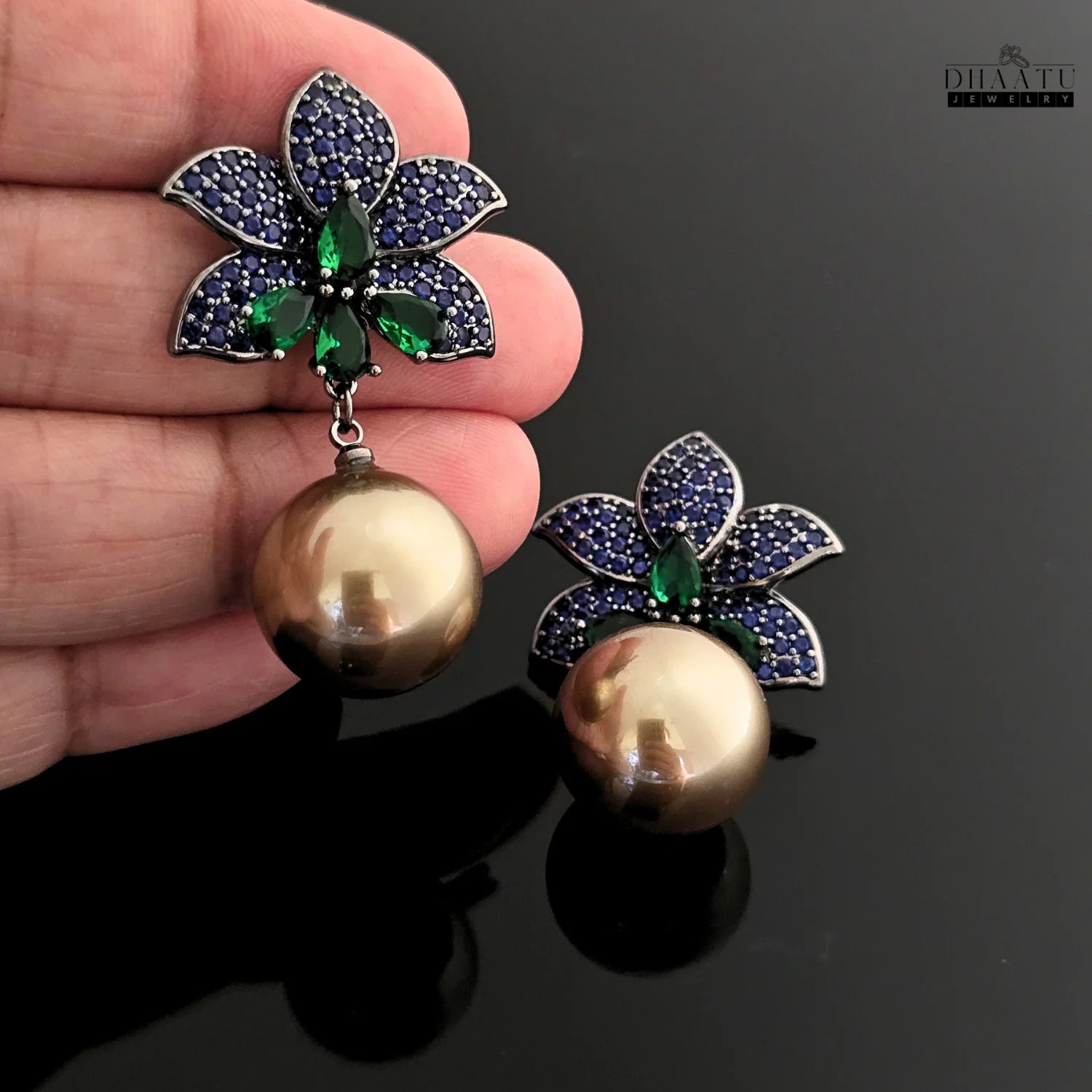 A hand holds one of the Dhaatu Jewelry blue sapphire and emerald flower earrings, showing the scale and detail of the pearl drop and floral stud.
