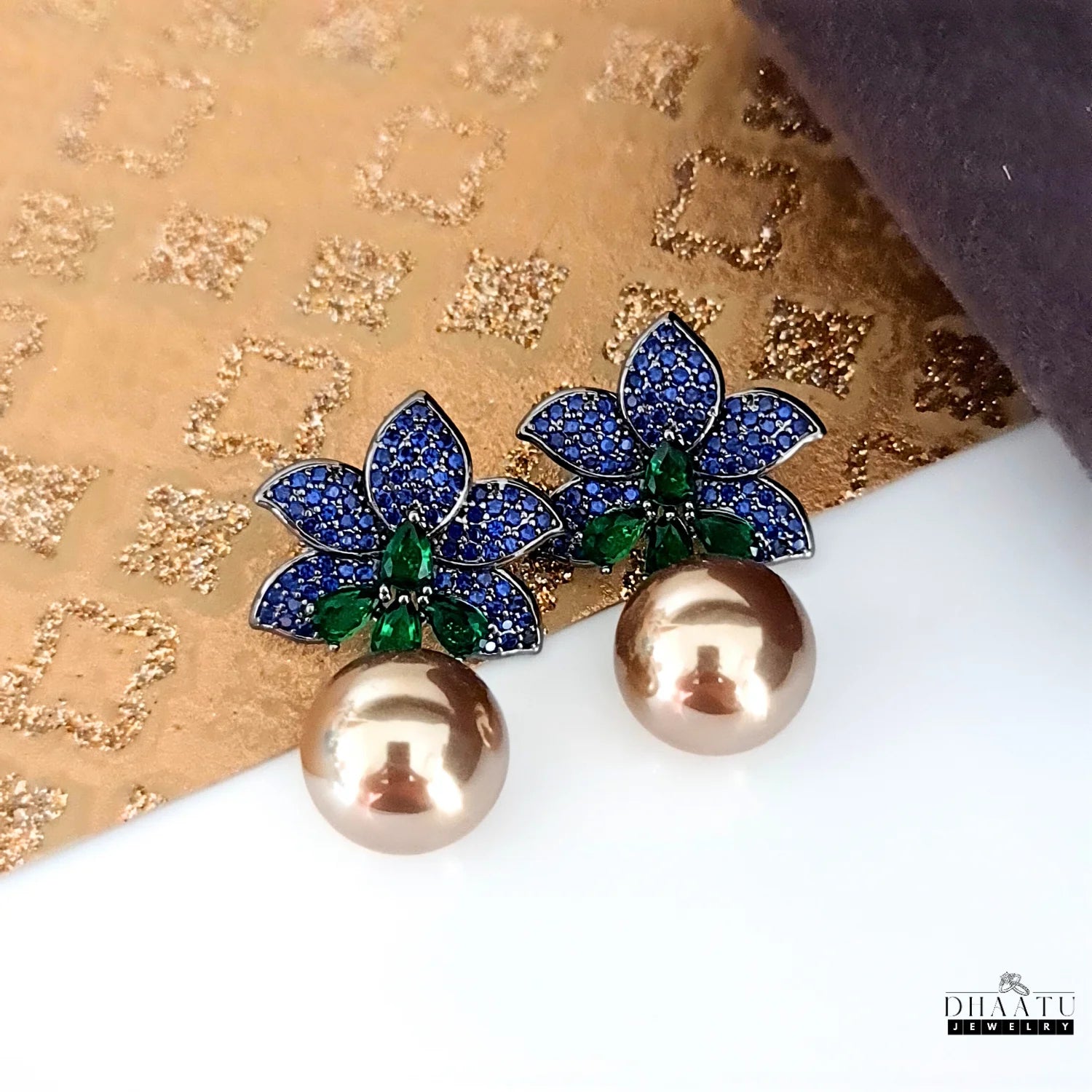 A close-up shot of the Dhaatu Jewelry blue sapphire and emerald flower earrings with a pearl drop, set against a shimmering gold background.