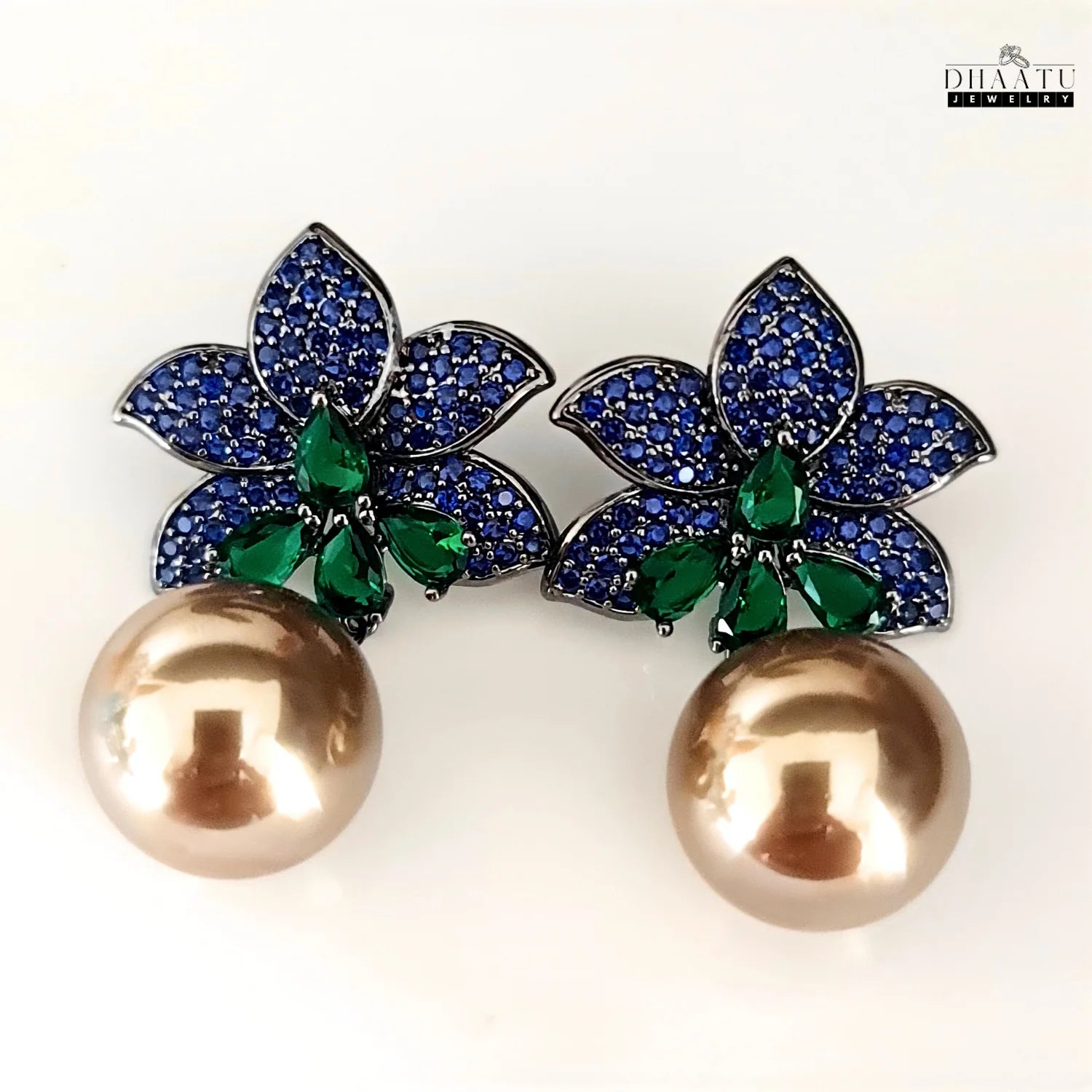 The Dhaatu Jewelry blue sapphire and emerald flower earrings with pearl drops shown on a clean white background, highlighting the contrast of the blue and green stones.