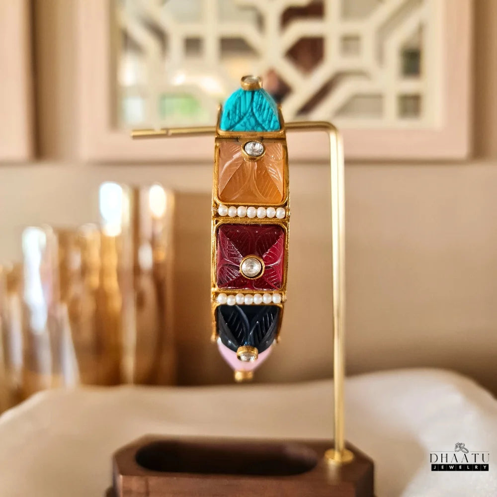 Angled view of a multicolor carved stone and pearl kada bracelet on a display stand, showcasing green, pink, blue, and red stones with gold plating.

