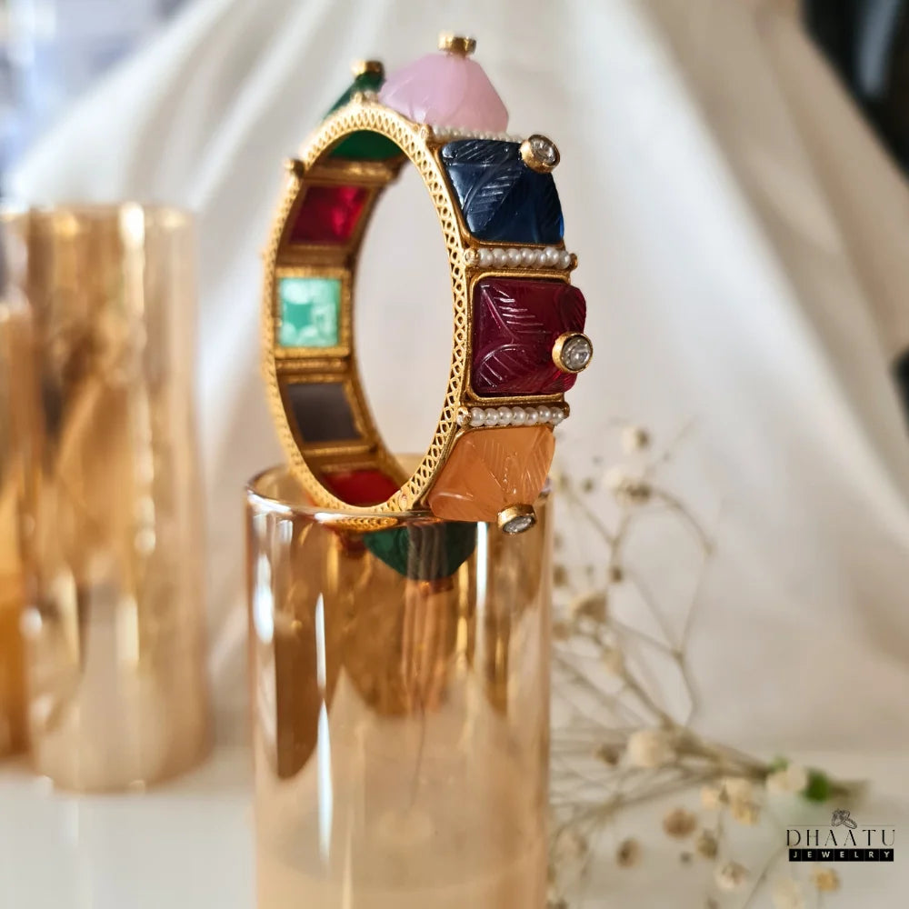 Top-down view of a multicolor carved stone and pearl kada bracelet on a white surface, featuring prominently green, pink, and black stones with pearl accents.

