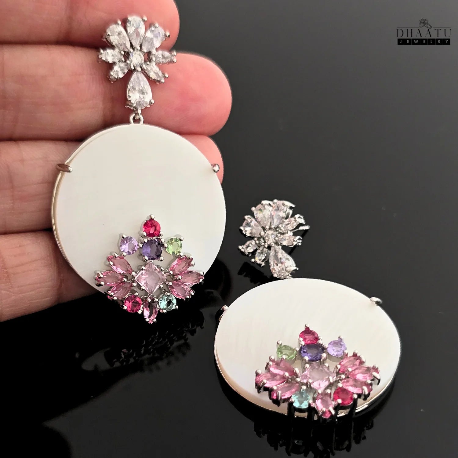 Hand holding Dhaatu Jewelry dangle earrings with floral stud and multi-colored stone accents.