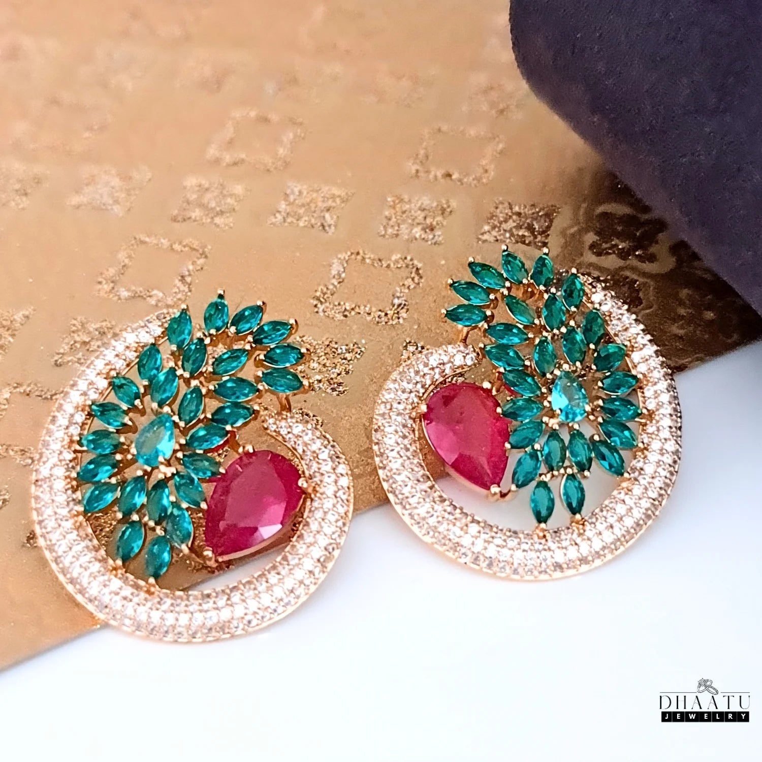 The Dhaatu green and ruby CZ stone stud earrings displayed on a subtle gold and white background, emphasizing their festive and luxurious aesthetic.