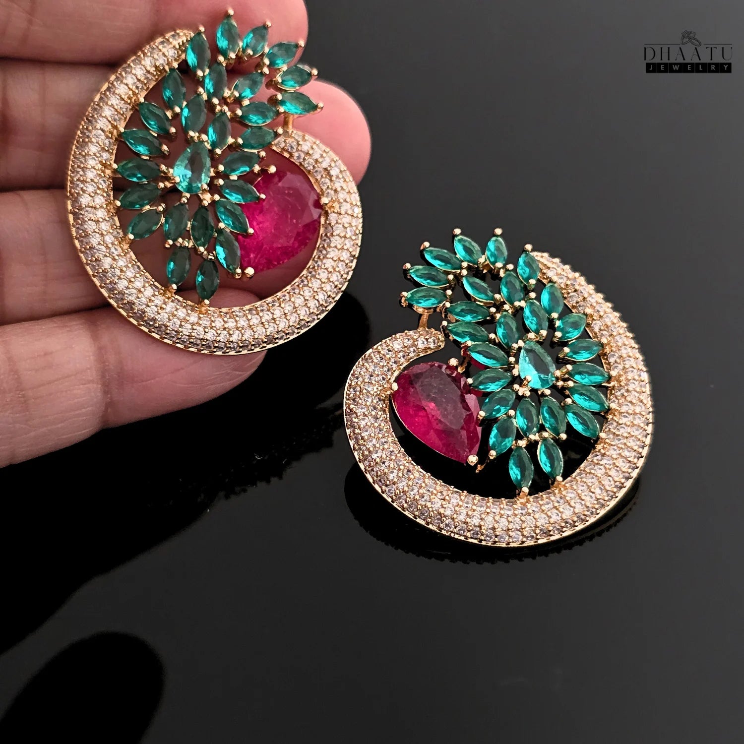 Close-up shot of the Dhaatu green and ruby CZ stud earrings held in a hand against a reflective black background, highlighting the gold plating and vibrant stones.