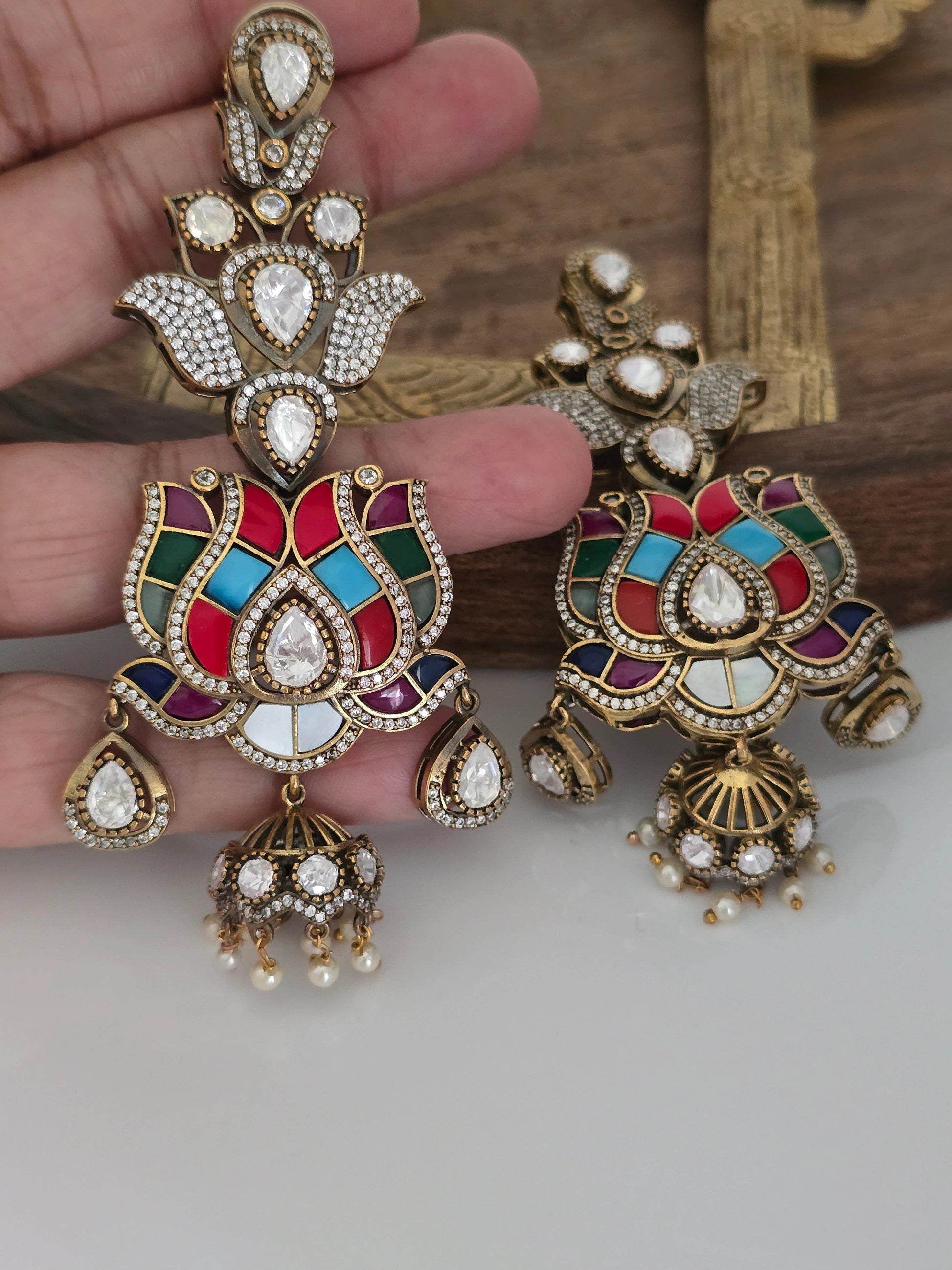 Pair of elaborate gold-tone lotus-shaped earrings with multicolored enamel (red, green, blue, turquoise, purple), set with polki-style American Diamonds and dangling jhumkas with small pearls, displayed on a beige and brown stand with one earring held in a hand.