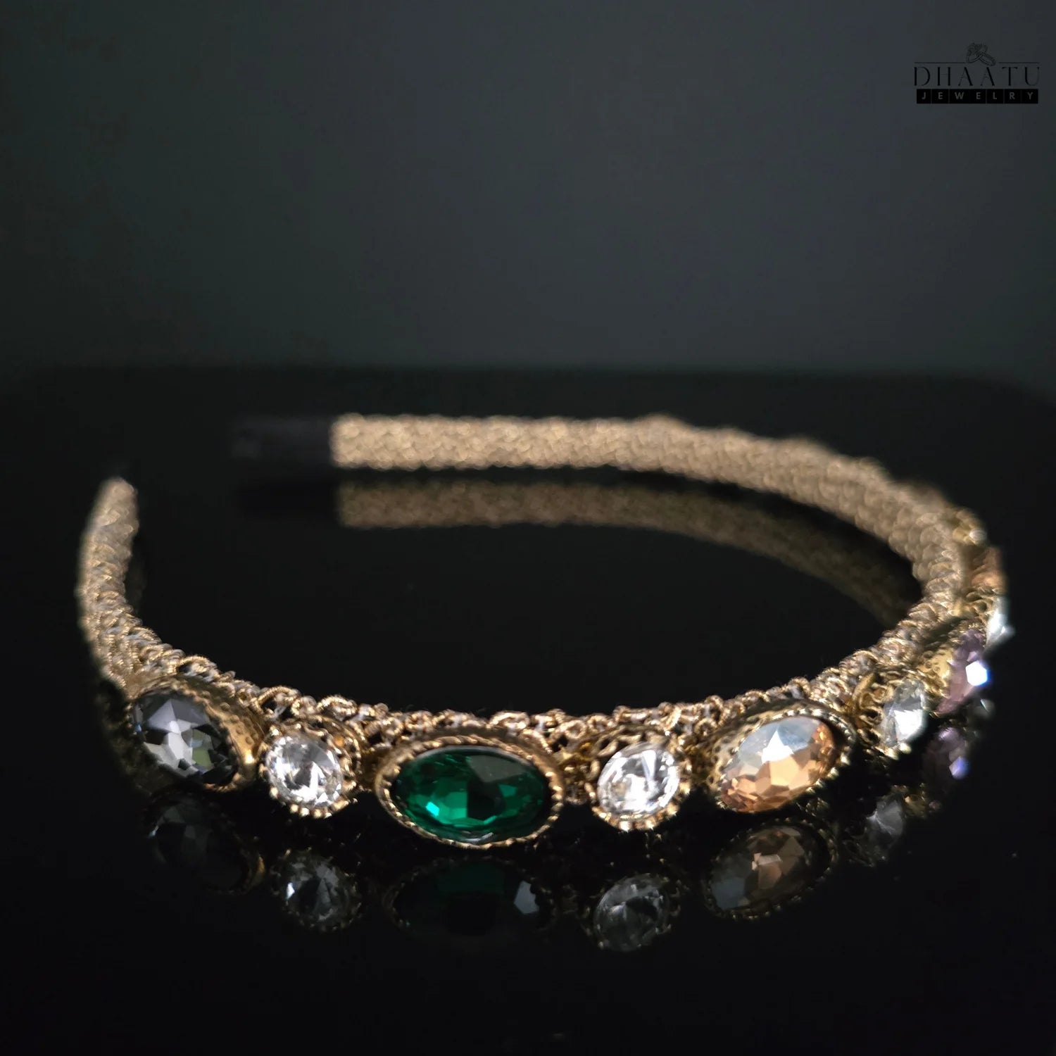 Close-up of Dhaatu Jeweled Headband showing gold-toned band with prominent green gemstone and clear, charcoal, and peach accent stones