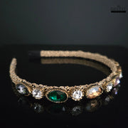 Close-up of Dhaatu Jeweled Headband showing gold-toned band with prominent green gemstone and clear, charcoal, and peach accent stones