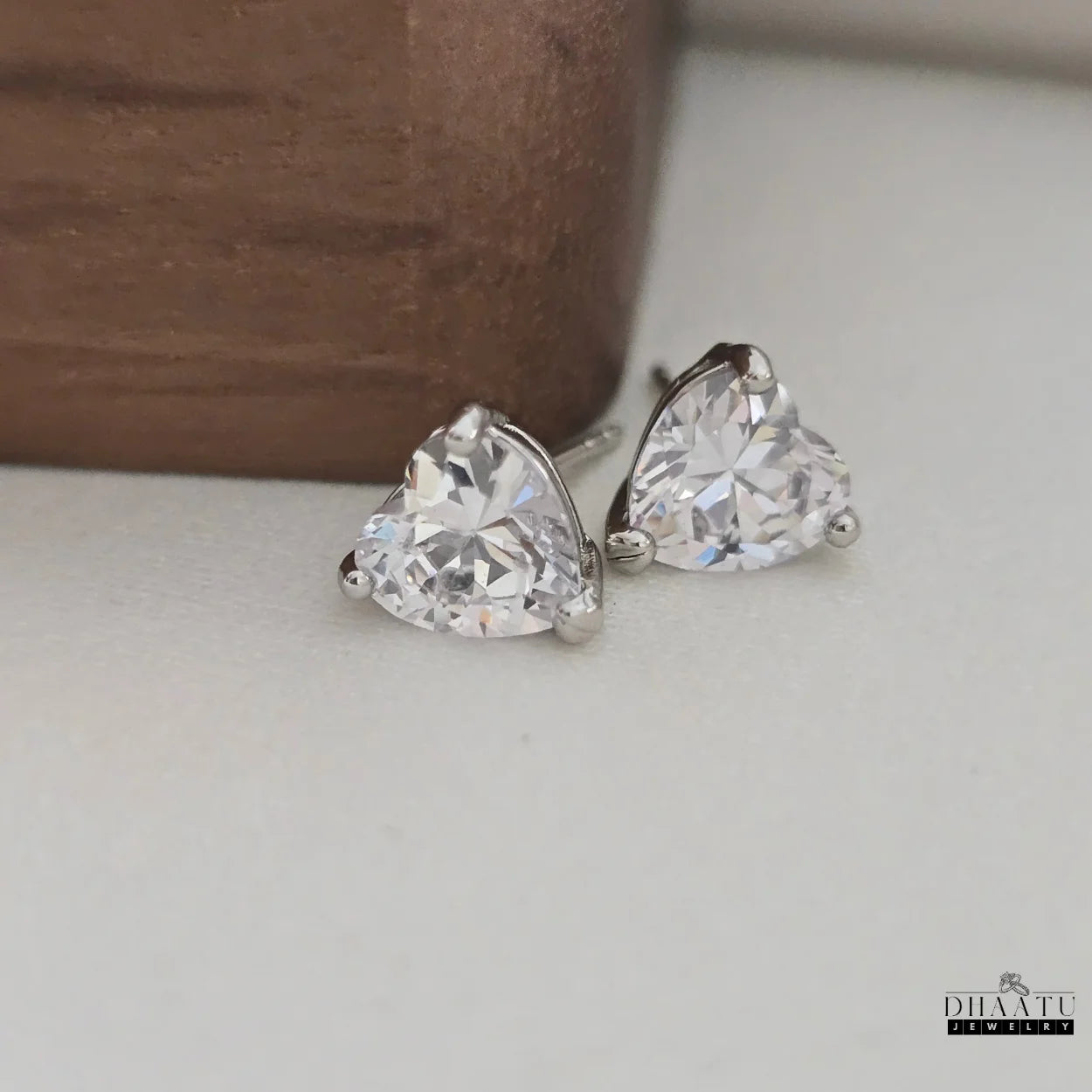 Close-up shot of two silver-tone heart-shaped AD stud earrings on a light background, showcasing their facets and brilliance