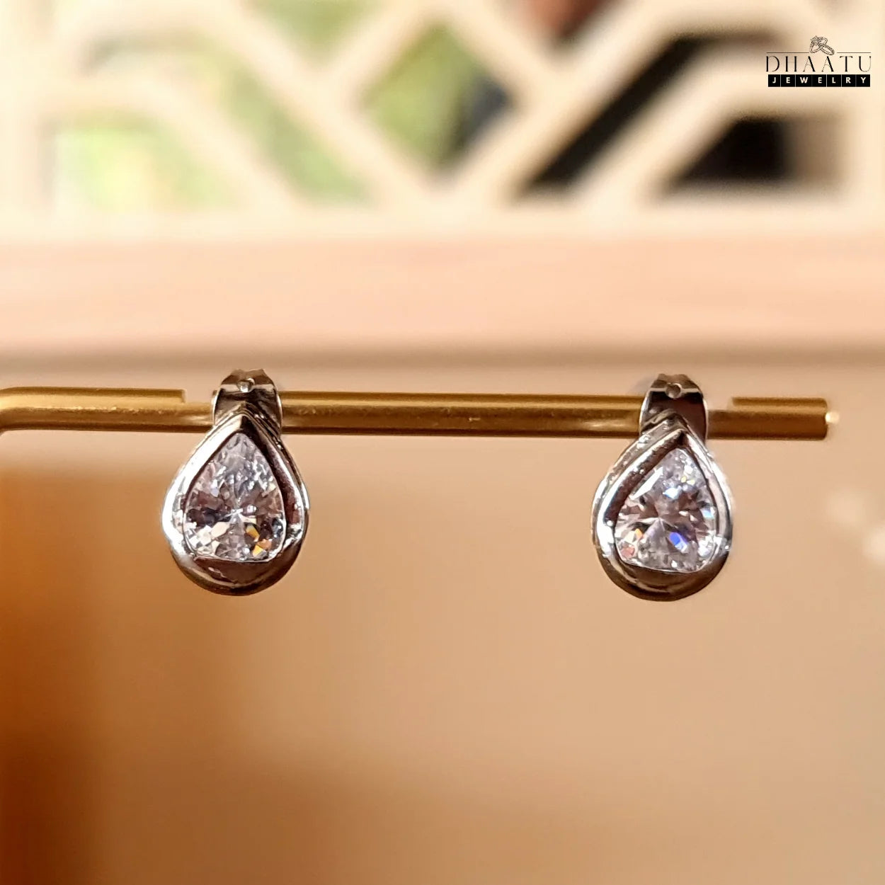 DHAATU JEWELRY pear-cut American diamond stud earrings hanging on a golden display stand, showcasing their rhodium-plated finish