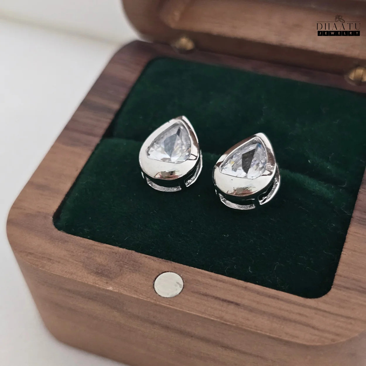 Pair of DHAATU JEWELRY pear-cut American diamond stud earrings resting in an open dark wooden jewelry box with green velvet interior.

