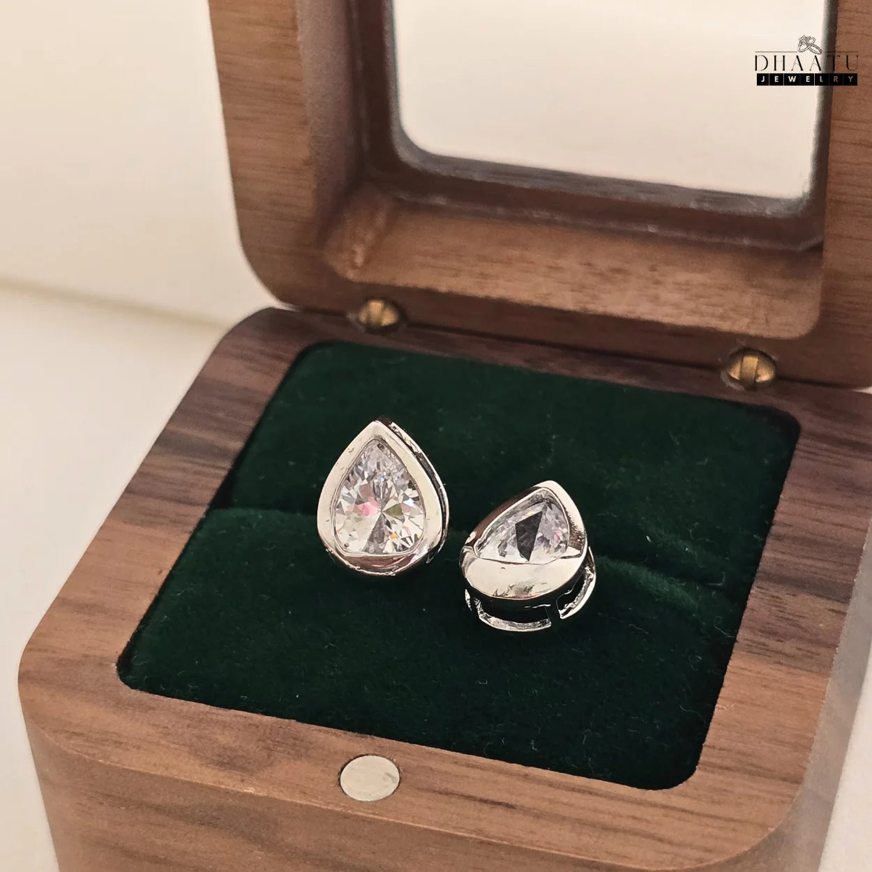 DHAATU JEWELRY pear-cut American diamond stud earrings displayed in a partially open wooden jewelry box with a clear lid.

