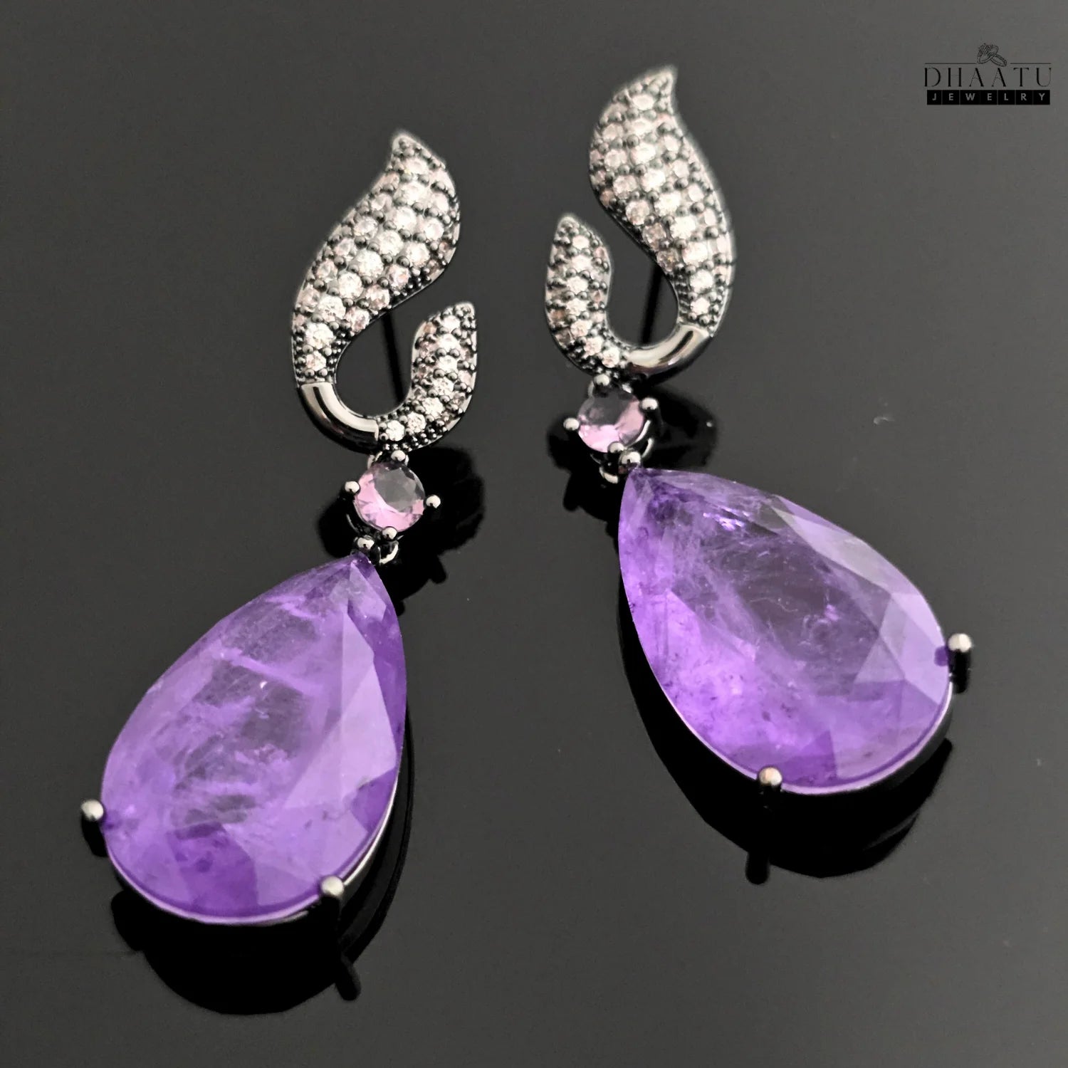 Dhaatu Jewelry dangle earrings with large purple amethyst stone and black rhodium plating on a reflective black surface.

