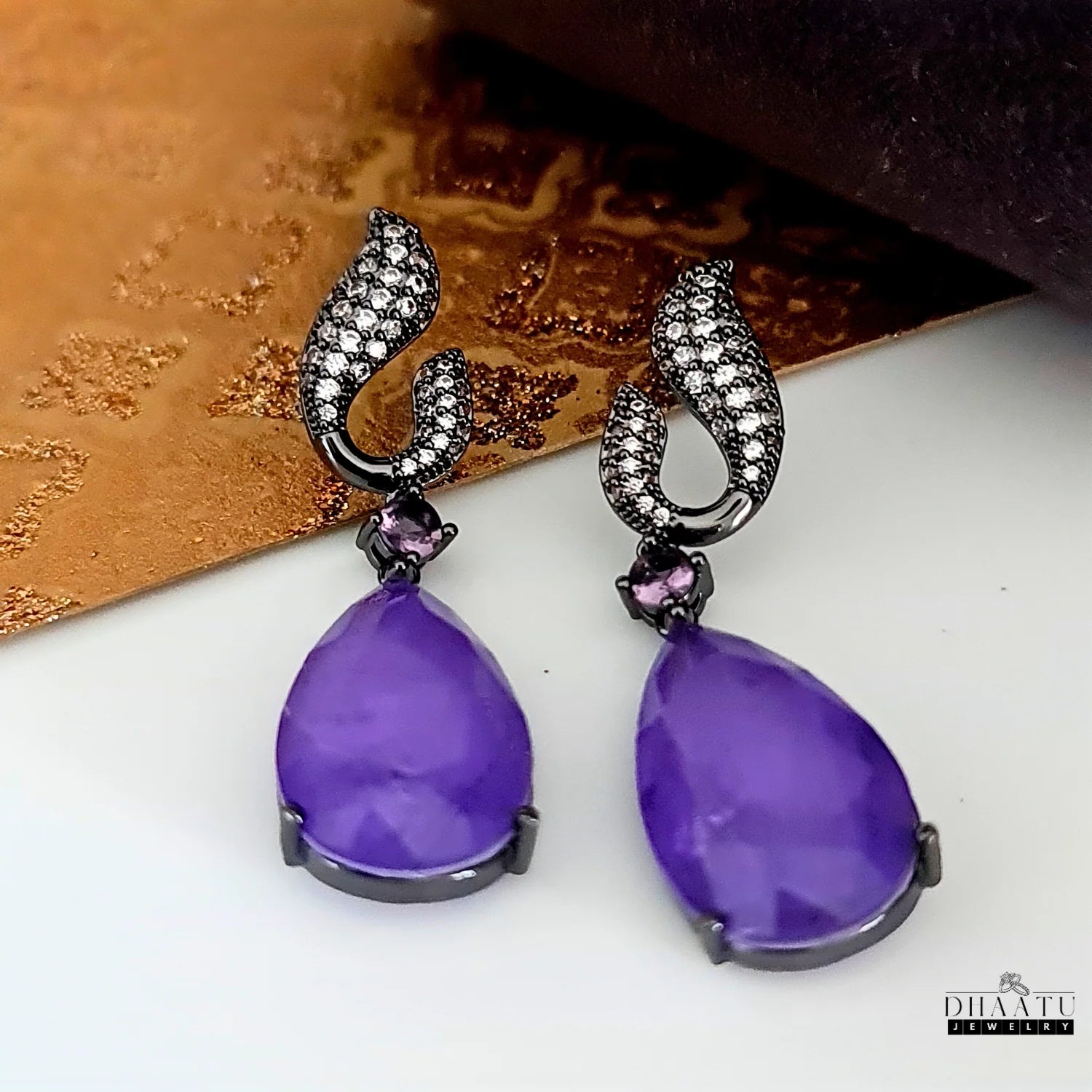 Close-up of Dhaatu Jewelry amethyst purple dangle earrings with black rhodium plating against a light background.


