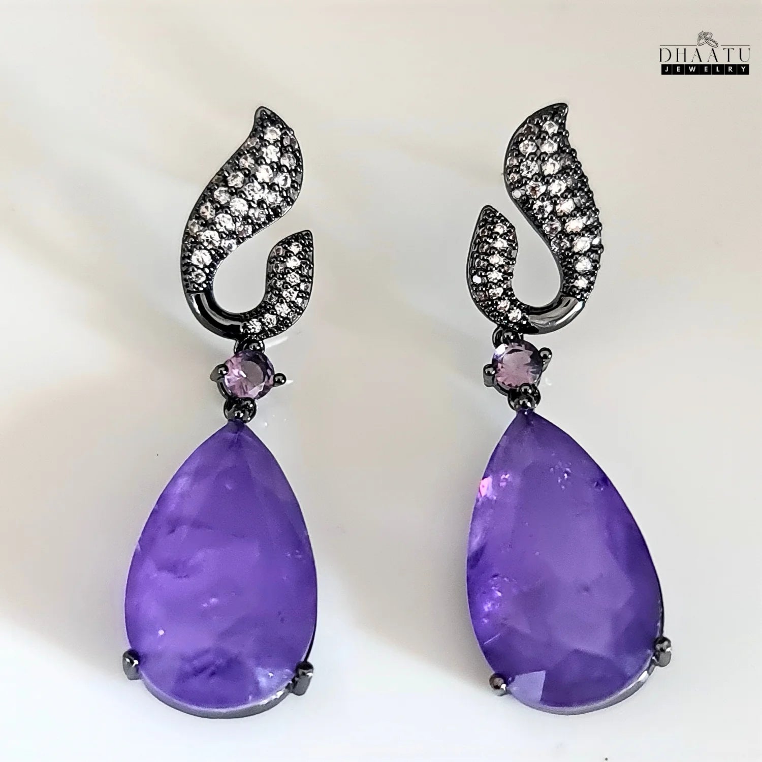 Close-up of Dhaatu Jewelry amethyst purple dangle earrings with black rhodium plating against a light background.

