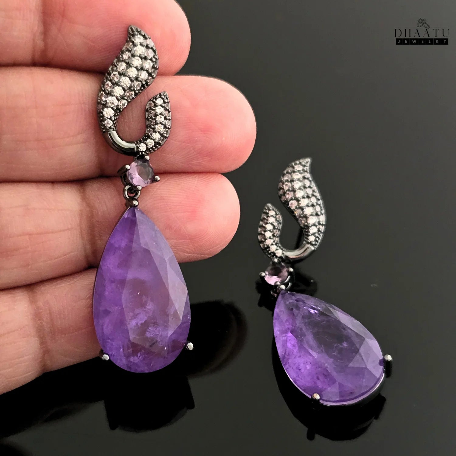 Hand holding up a Dhaatu Jewelry amethyst purple stone dangle earring, highlighting the black rhodium plating and stone size.

