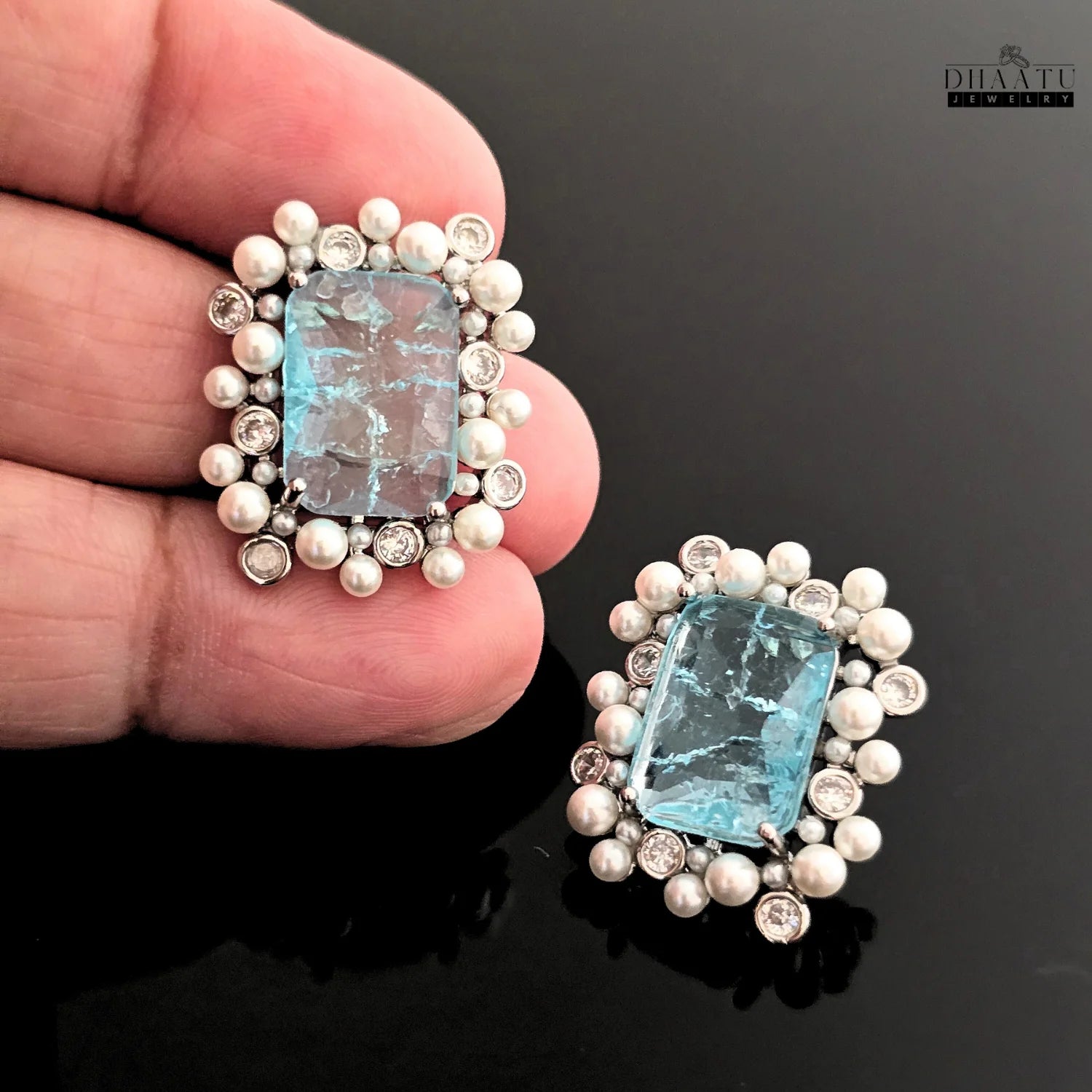 Pair of earrings with blue gemstones and pearl-like beads held by a hand on a dark background.