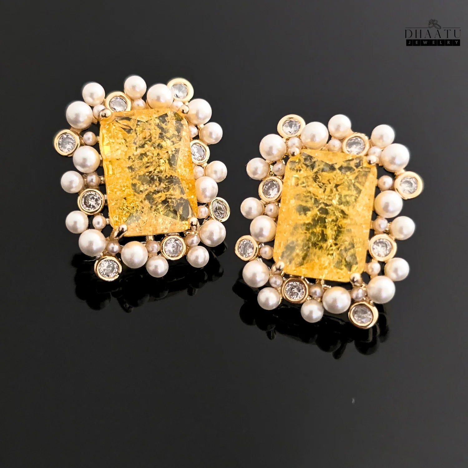 Closeup of DHAATU JEWELRY stud earrings featuring a rectangular citrine stone with gold flakes, framed by polki and pearls on a black reflective surface.

