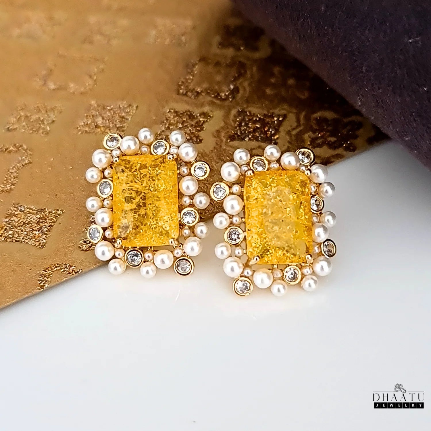 Product shot of DHAATU JEWELRY studs on a mixed gold and white textured background, highlighting the golden hues of the earrings.