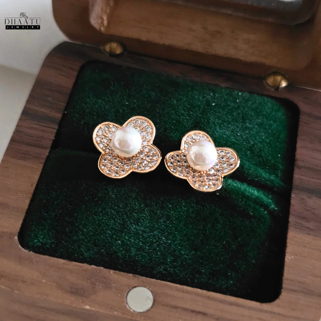 DHAATU JEWELRY gold-plated clover pearl and zircon stud earrings resting on green velvet inside a wooden jewelry box.

