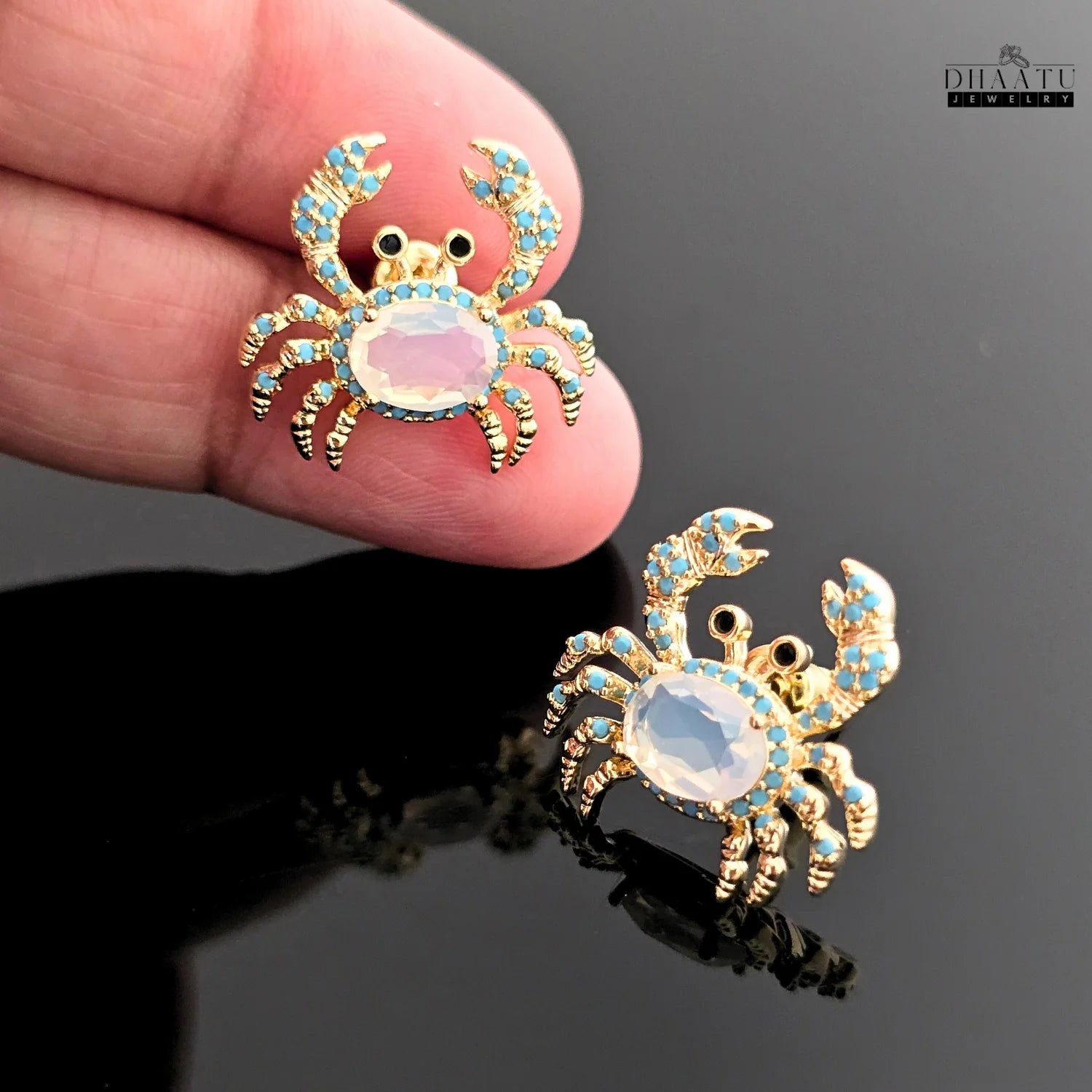 Closeup of a pair of gold-toned crab earrings with turquoise and white zircon stones, held in a hand.