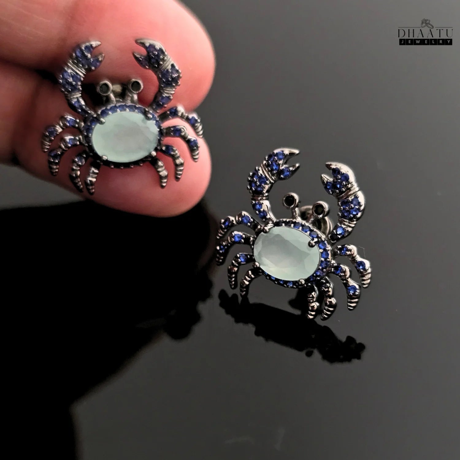 Closeup of a pair of silver-toned crab earrings with dark blue and aquamarine zircon stones, held in a hand.