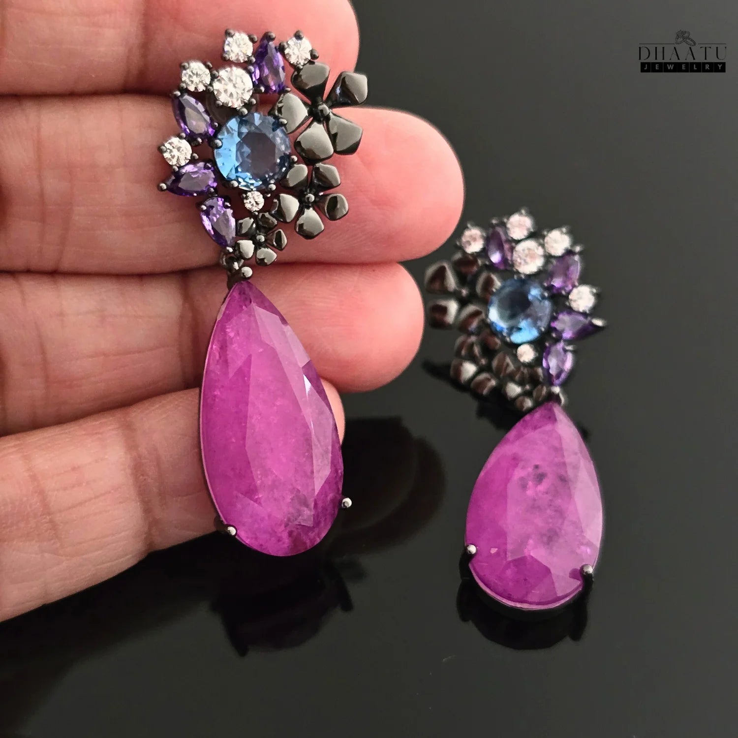 Close-up of DHAATU JEWELRY drop earrings held in a hand to show size and scale, highlighting the detailed craftsmanship and violet teardrop stone.
