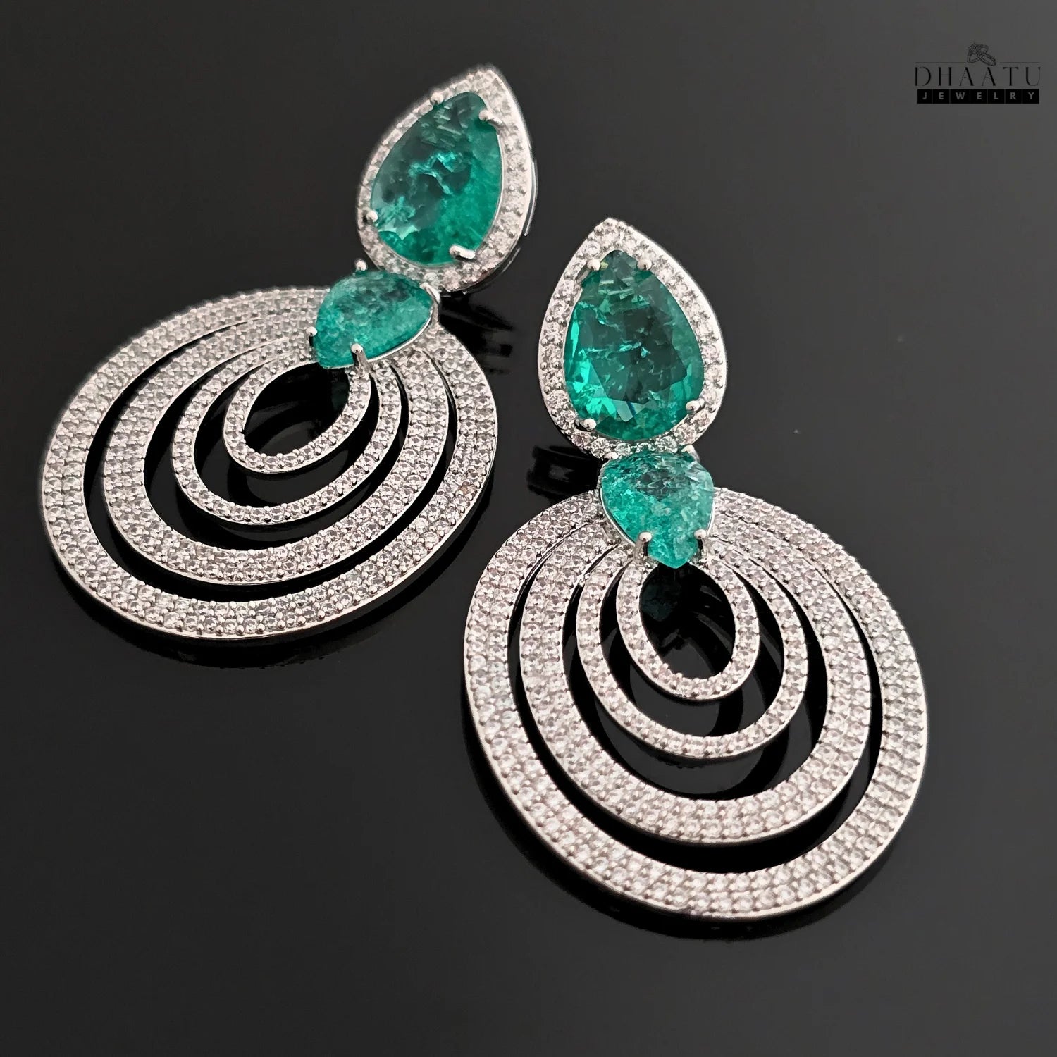 A pair of DHAATU JEWELRY emerald green and pave diamond chandelier earrings, shown at an angle on a black reflective surface.